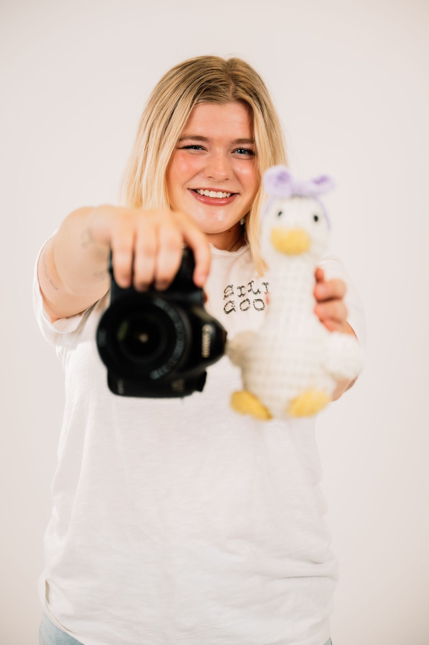 Smiling young blonde holding DSLR camera and duck plush toy toward camera in bright white studio.