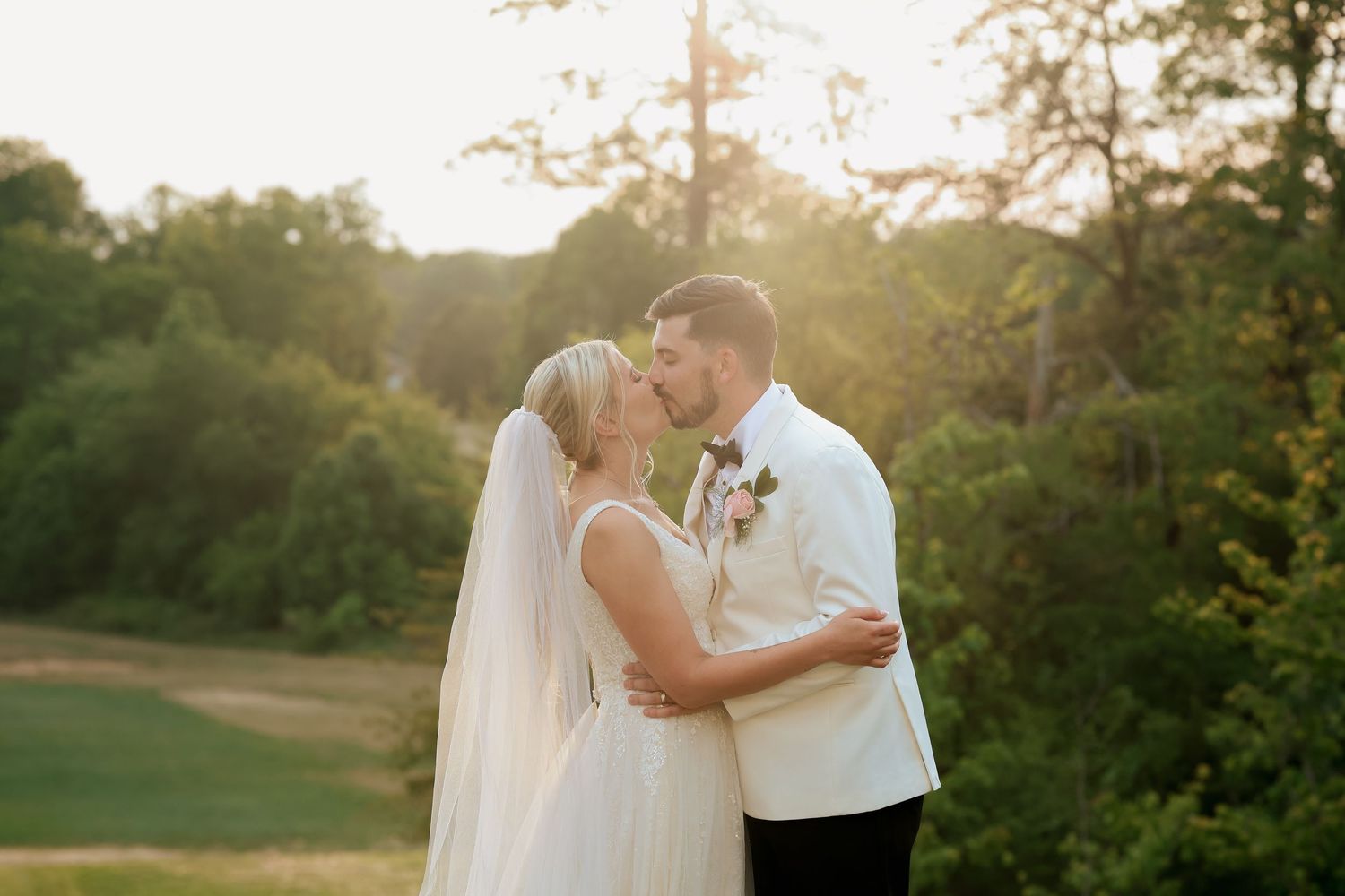 Newlyweds share a golden hour kiss outdoors surrounded by lush green trees at wedding.