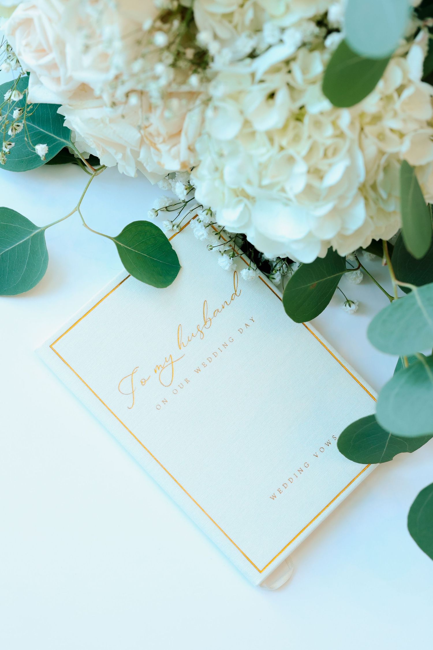Wedding vow book addressed to husband with gold border beside white hydrangea bouquet.