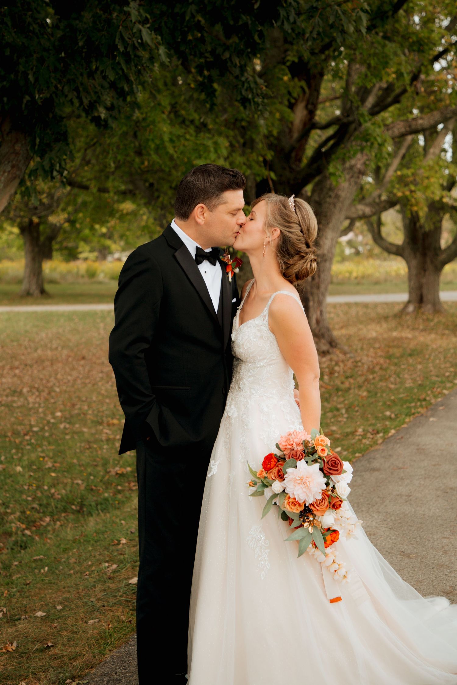 Bride and groom kissing while holding cascading autumn floral bouquet under park trees.