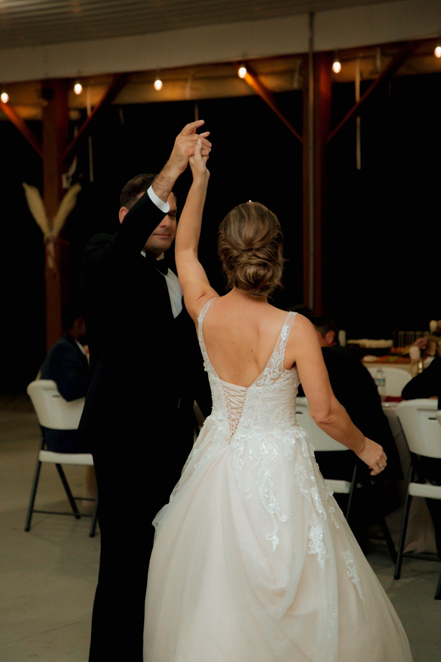 Groom spins bride in elegant lace wedding gown during first dance at barn reception.
