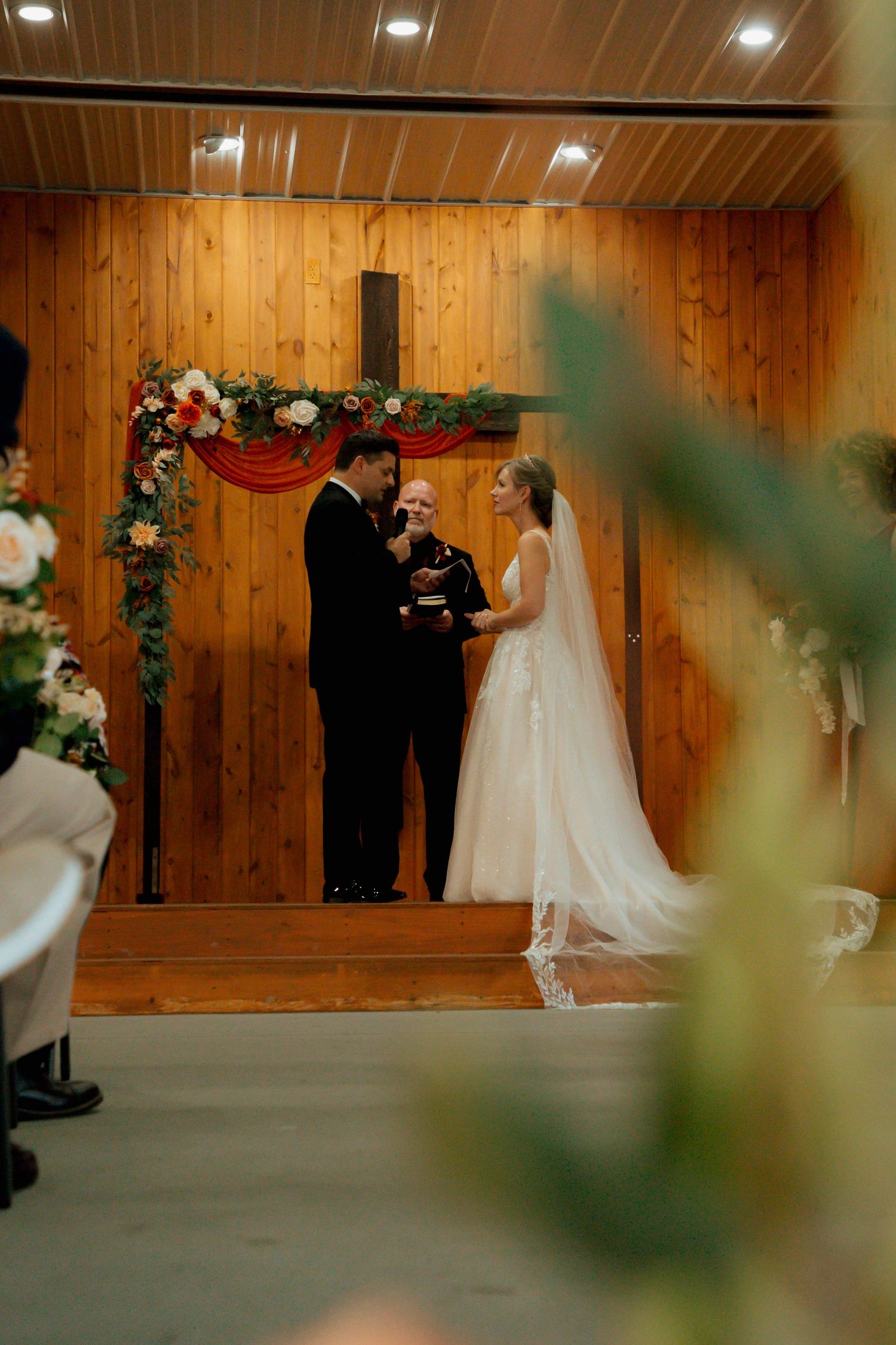 Bride and groom hold hands before officiant at rustic chapel altar with floral cross arch.