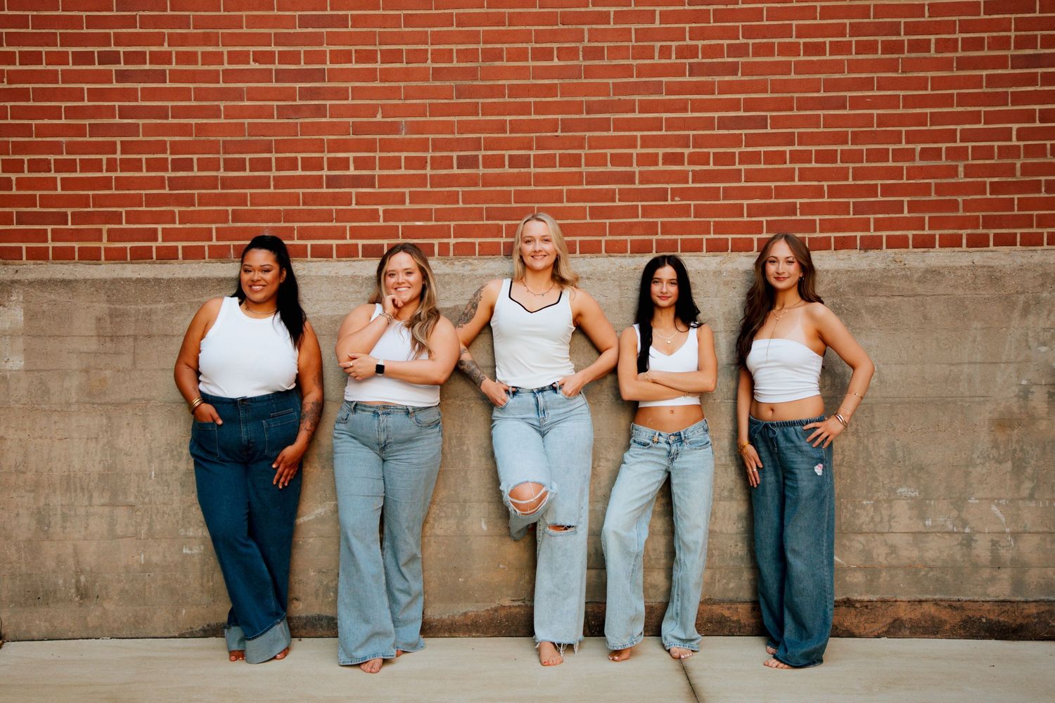 Five women wearing white crop tops and jeans posing confidently against a red brick wall outdoors.