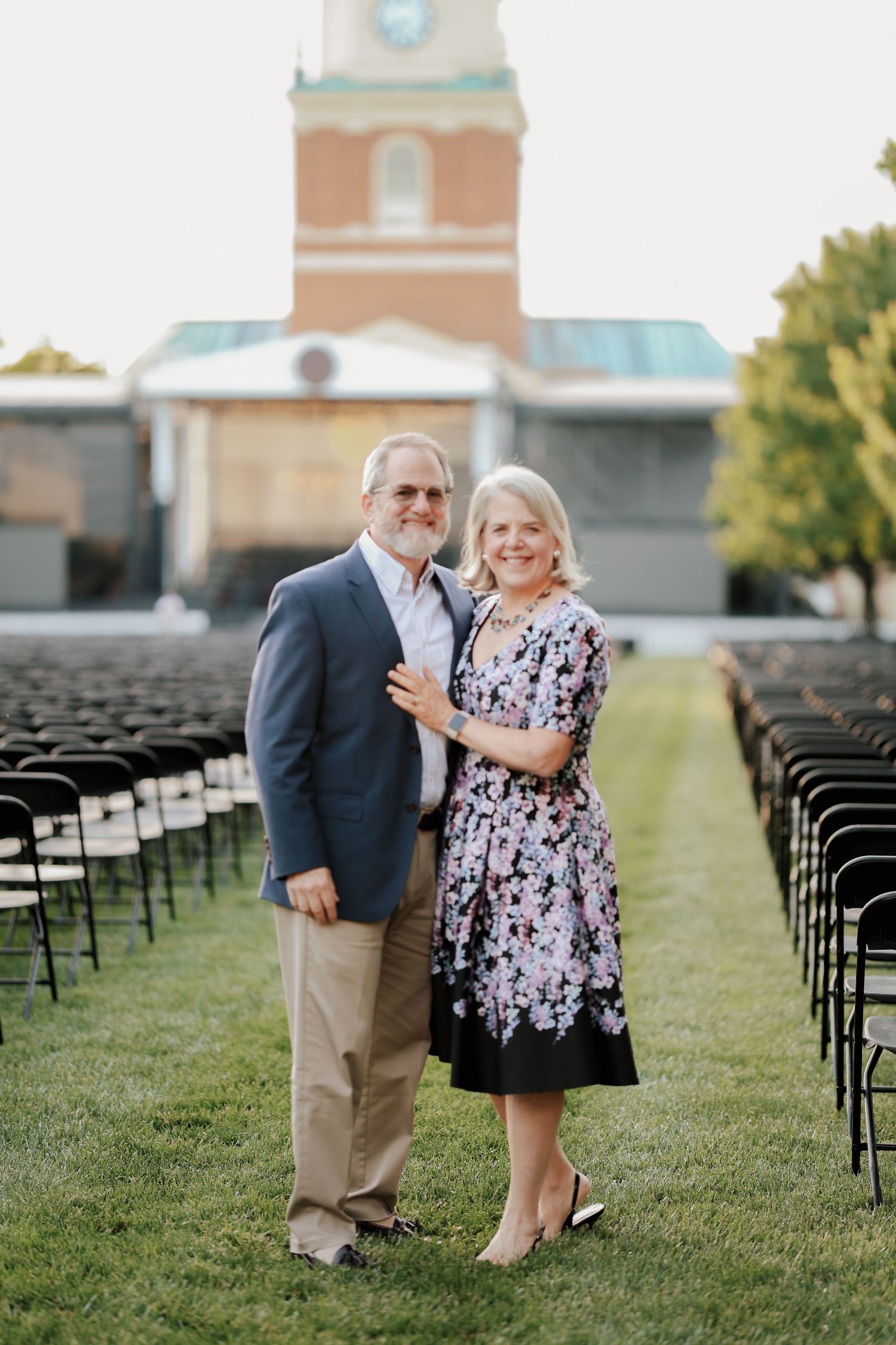 Couple posing together on green lawn with rows of black chairs and brick church building behind them.