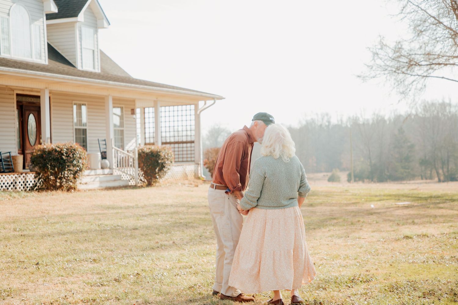 Elderly couple sharing an intimate embrace on a sunlit lawn in front of a classic white farmhouse.