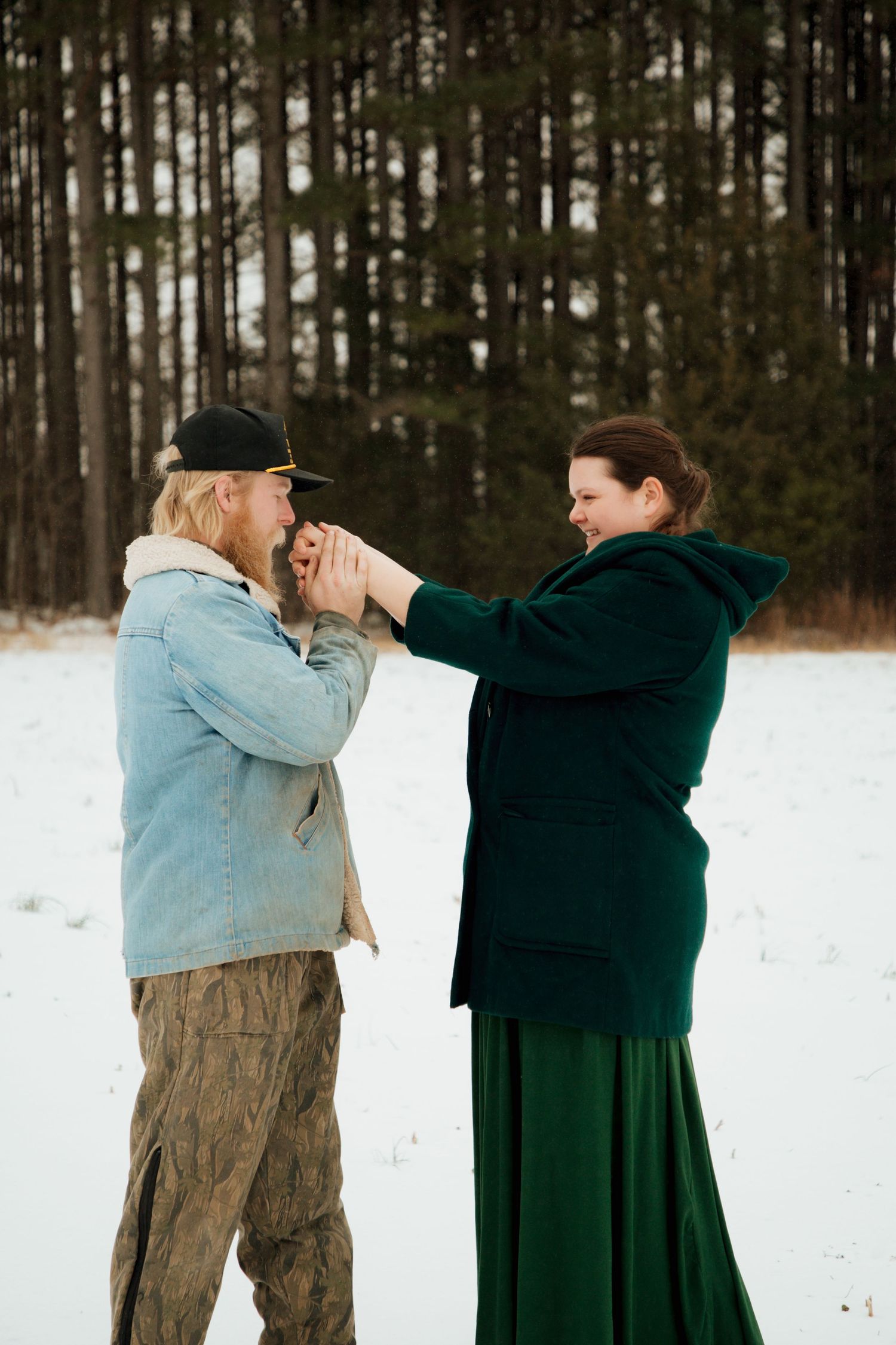 Couple warming hands together in snowy field with pine trees, woman in green velvet coat.