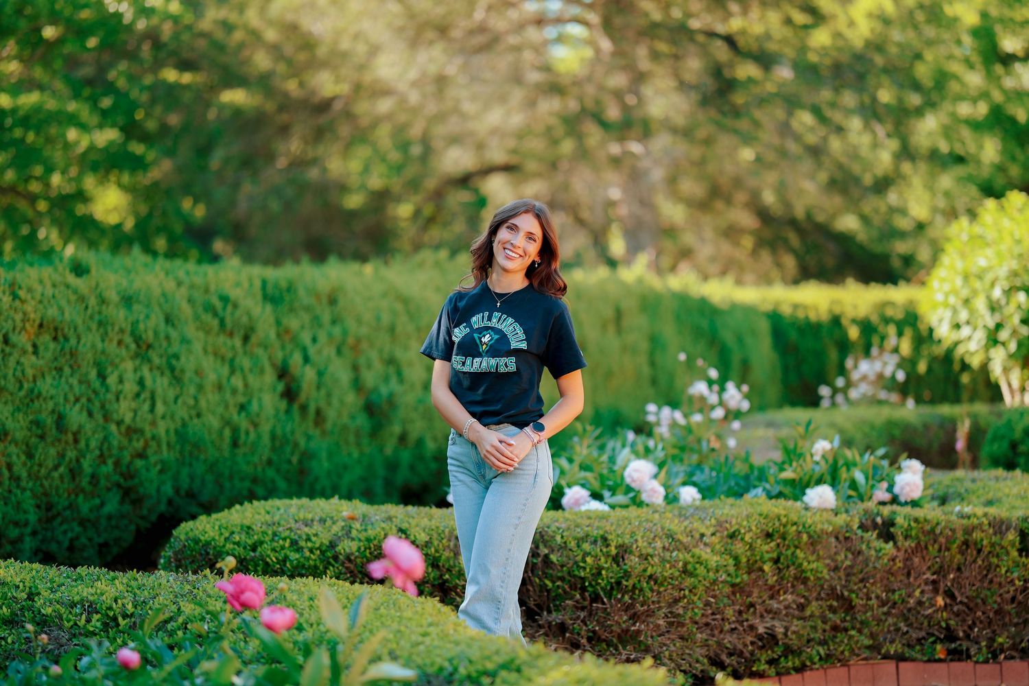 Young woman in dark college t-shirt poses in lush garden with trimmed hedges.