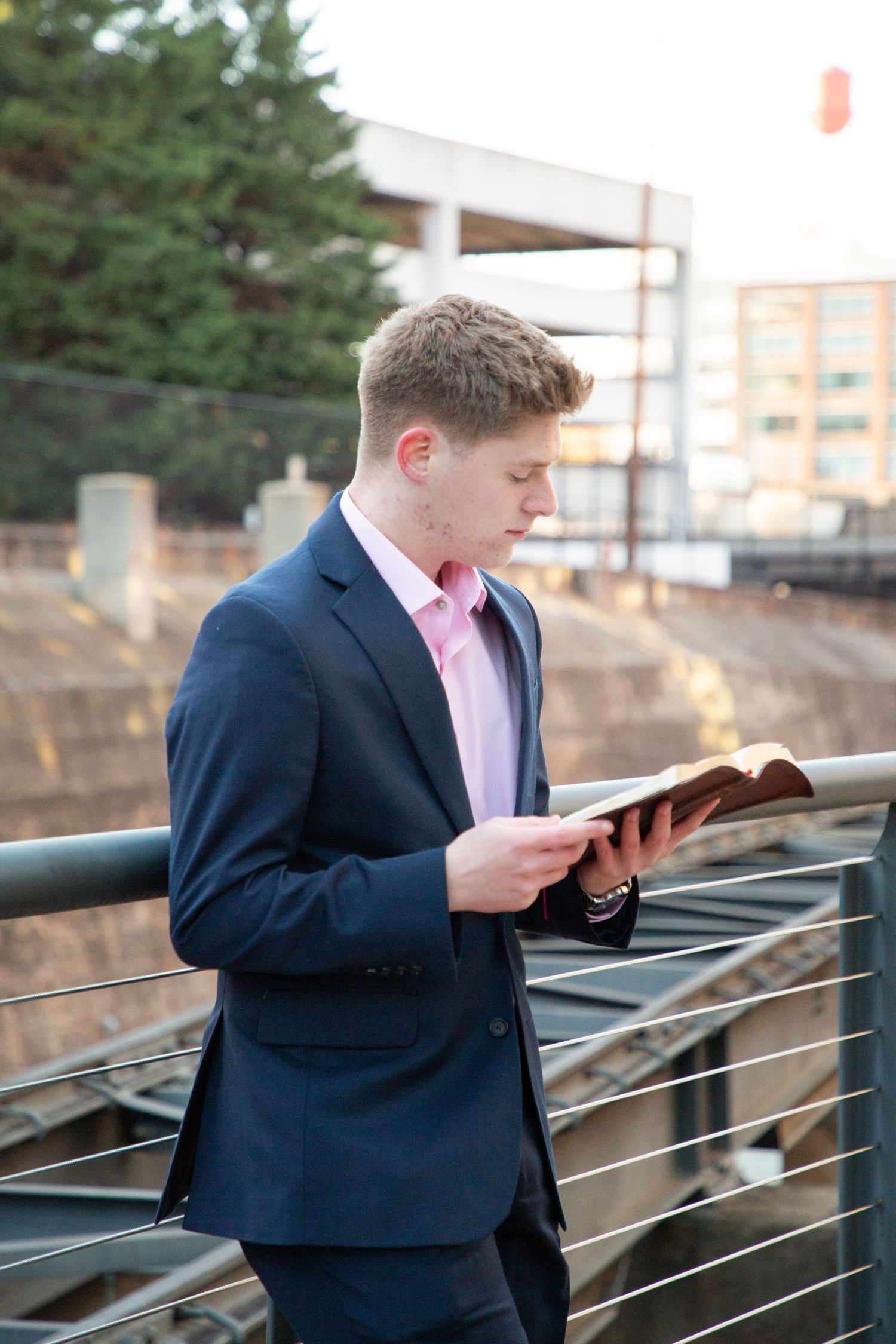 Young man in navy suit and pink shirt reading a book on an urban bridge.