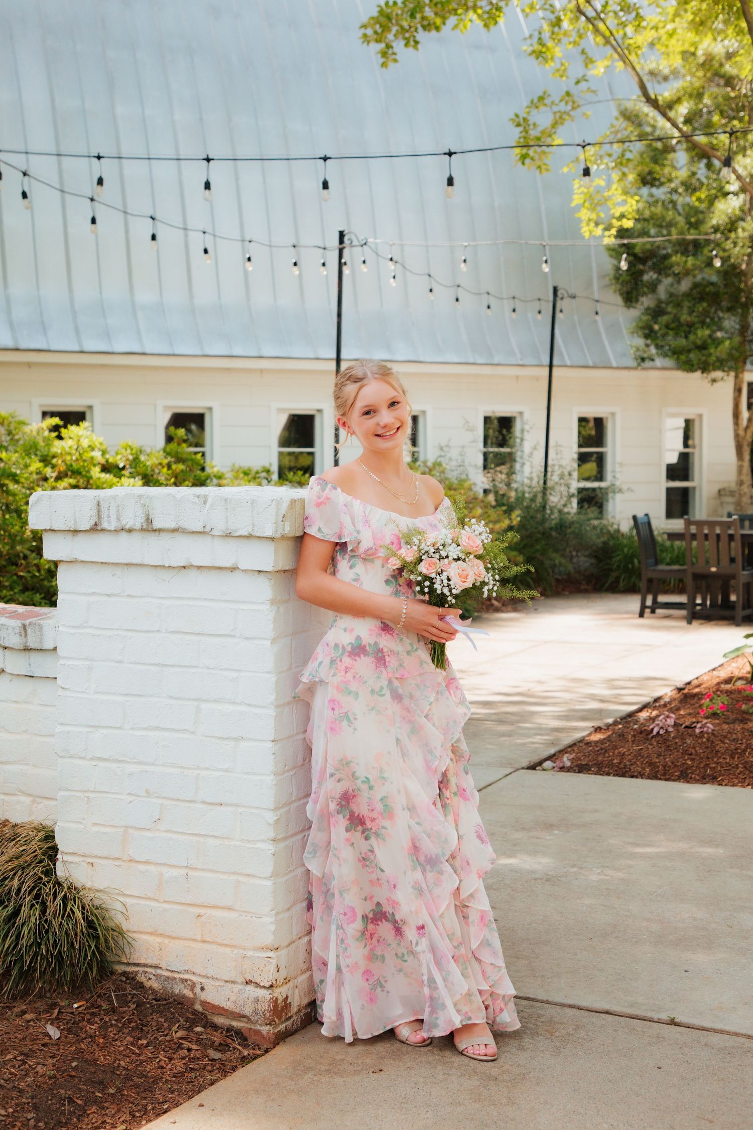 Smiling young woman in pink floral off-shoulder ruffle gown holding bouquet, leaning against white brick wall outdoors.