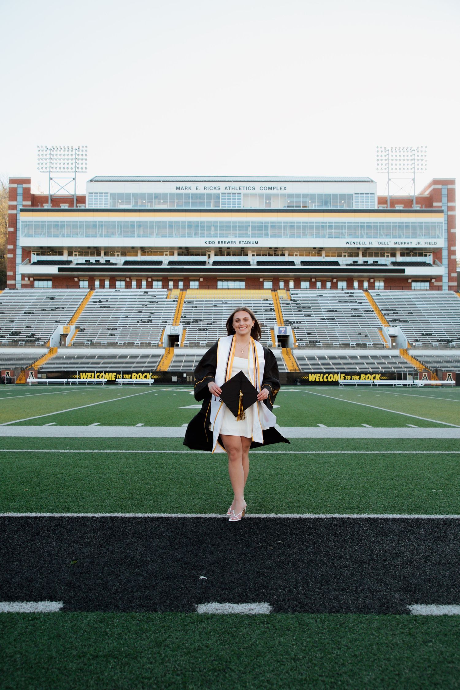 Graduate in cap and gown walking on Kidd Brewer Stadium field at Appalachian State University.