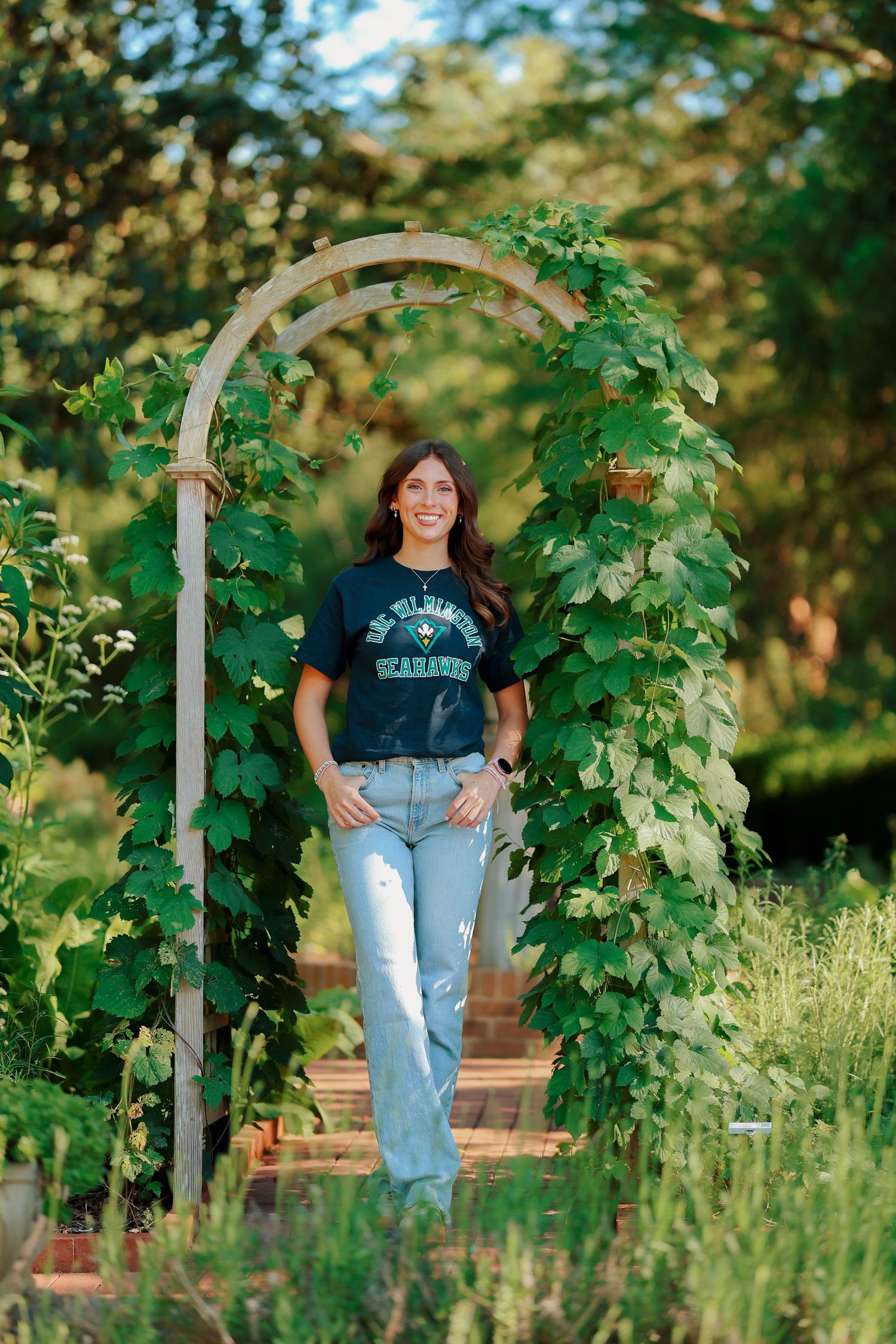 Smiling young woman in UNCW Seahawks tee standing under a vine-covered wooden garden arch.