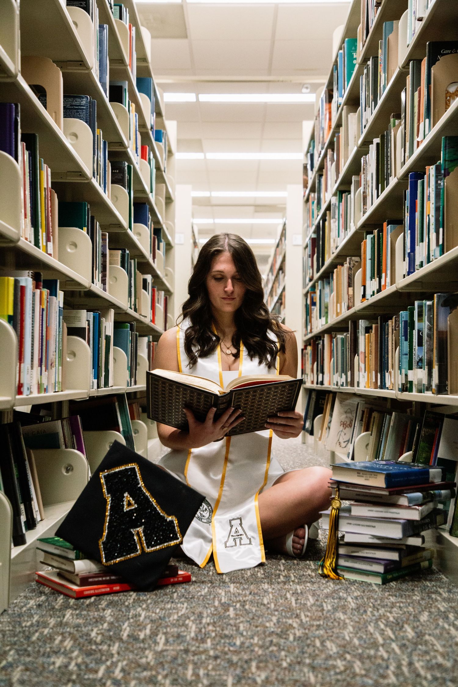 Graduate sitting on library floor reading book surrounded by stacked books and cap.