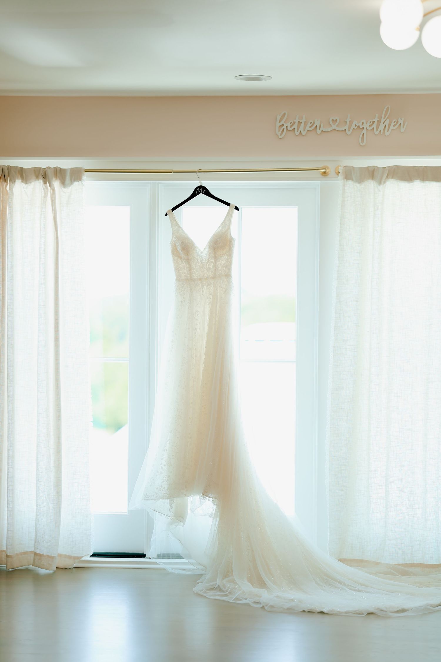 Elegant white lace wedding dress hanging in window with better together sign above curtains.
