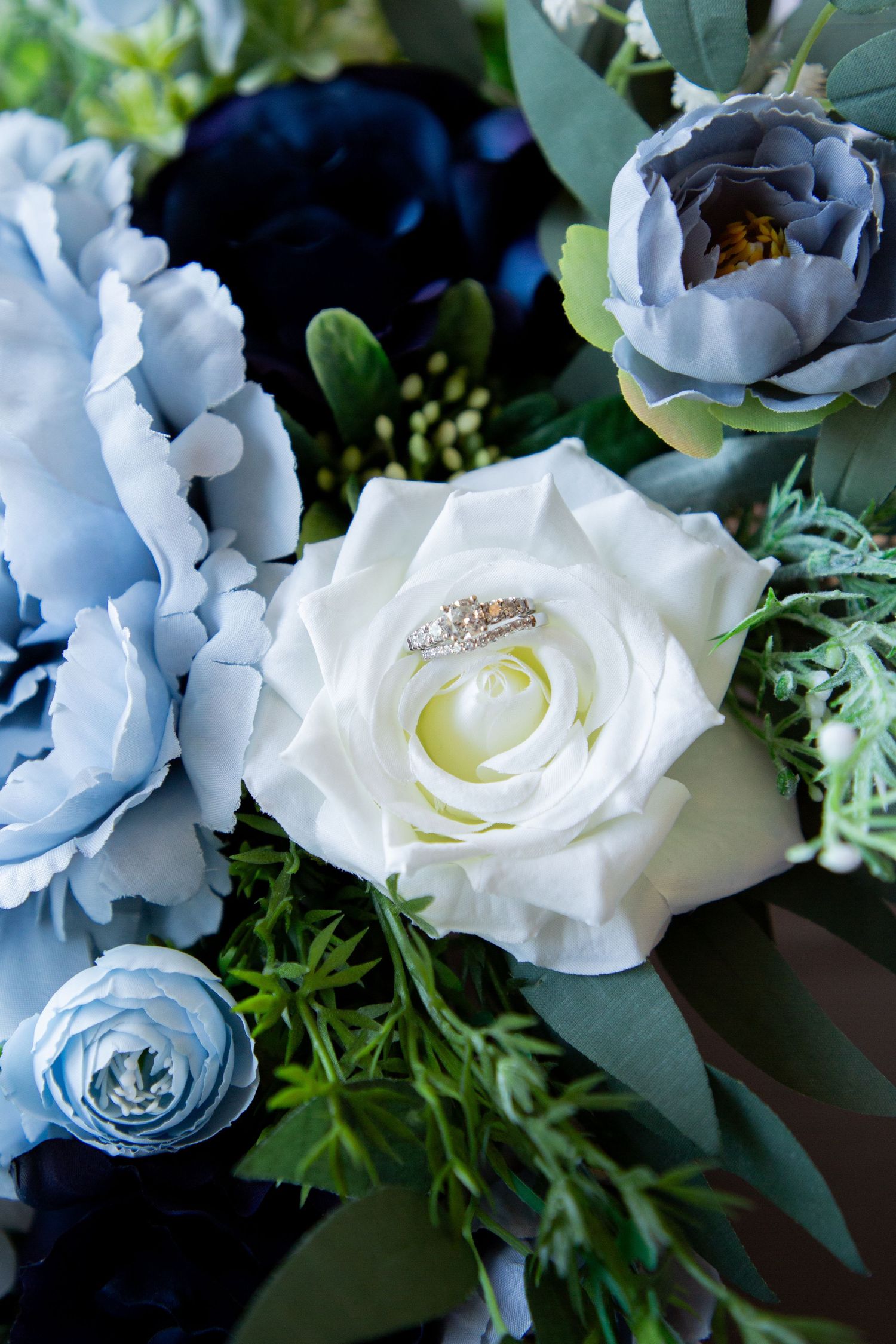 Diamond wedding ring placed inside a white rose surrounded by blue and navy flowers in a bouquet.