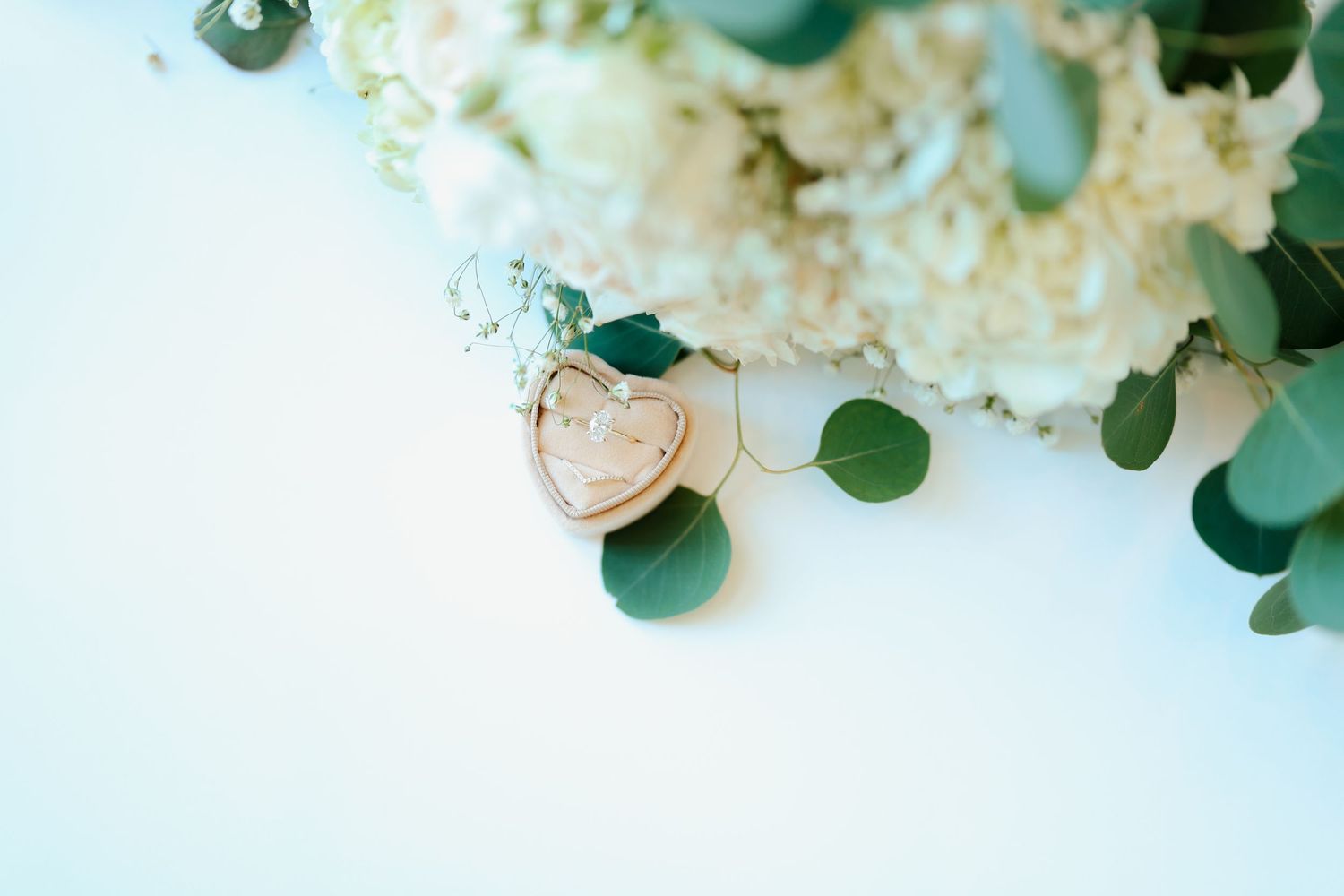 Heart-shaped wooden ring box rests near white hydrangea and eucalyptus wedding bouquet on a light background.