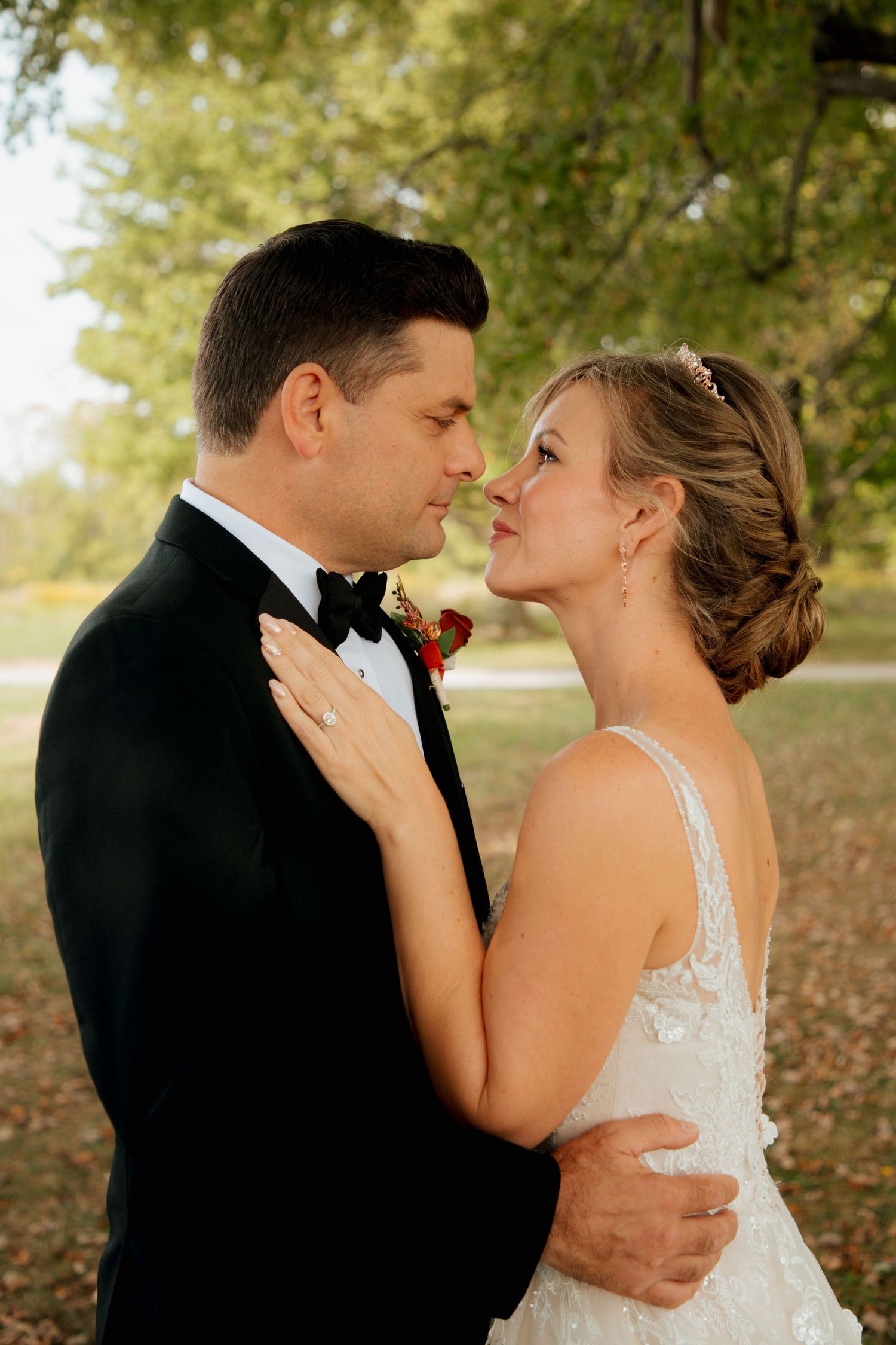 Bride and groom share a romantic moment, nearly kissing outdoors with lush green trees in the background.