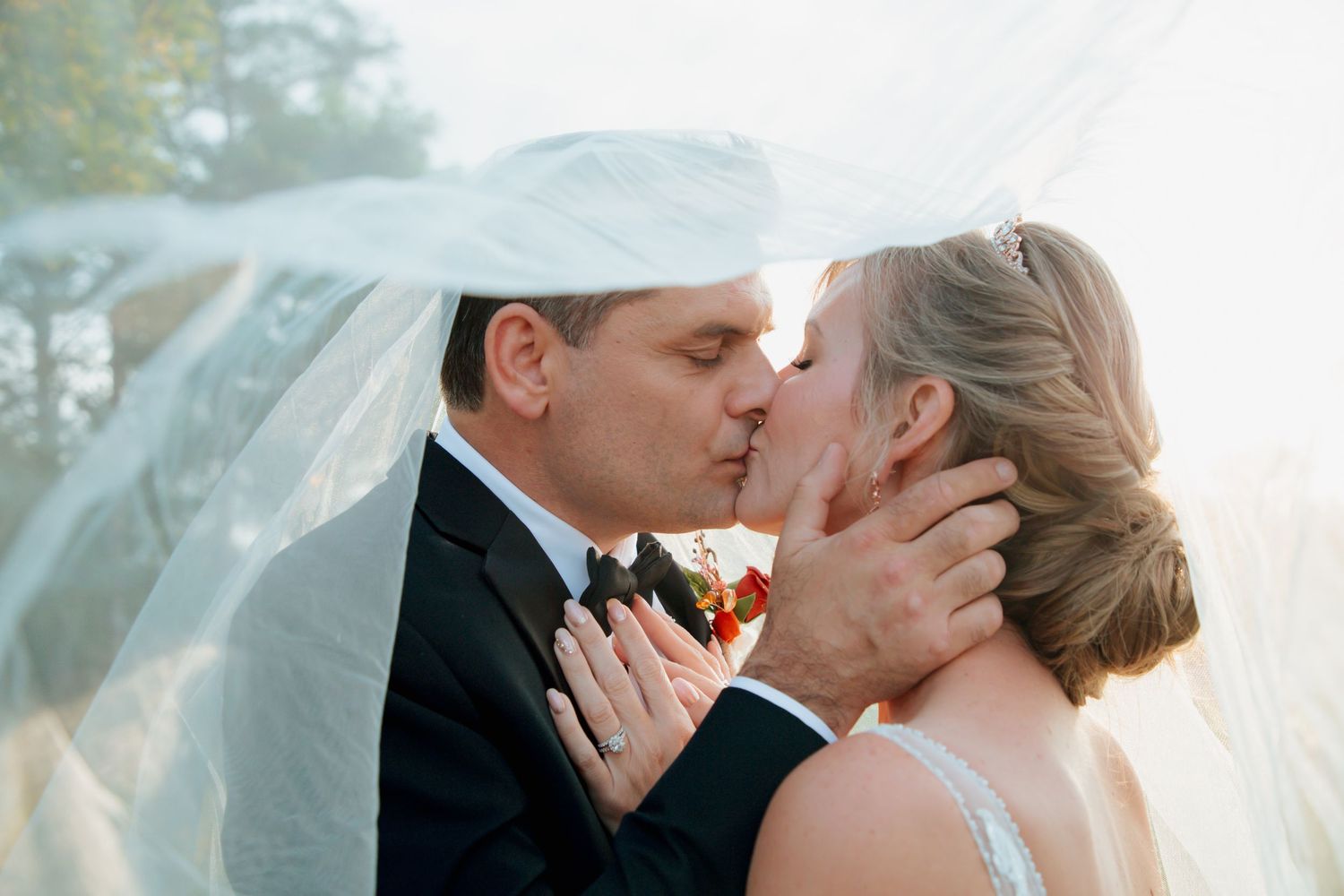 Bride and groom share a tender kiss beneath the bridal veil in soft golden outdoor wedding portrait.