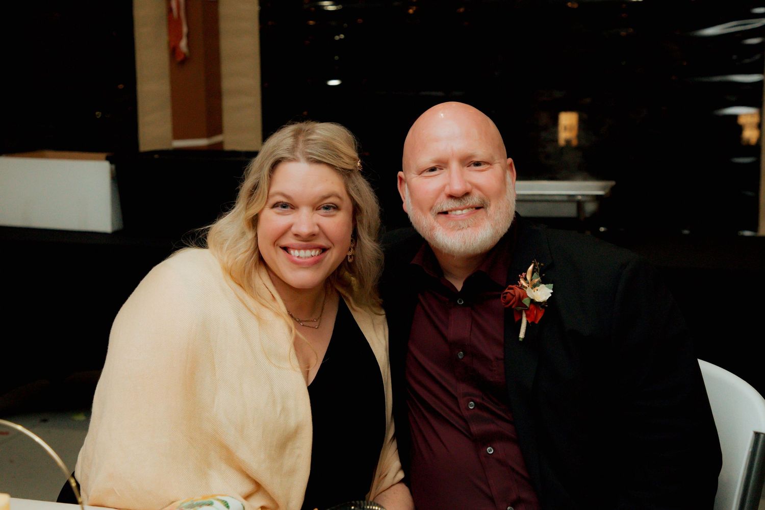 Smiling couple posing together at a wedding reception, man wearing boutonniere in black suit.