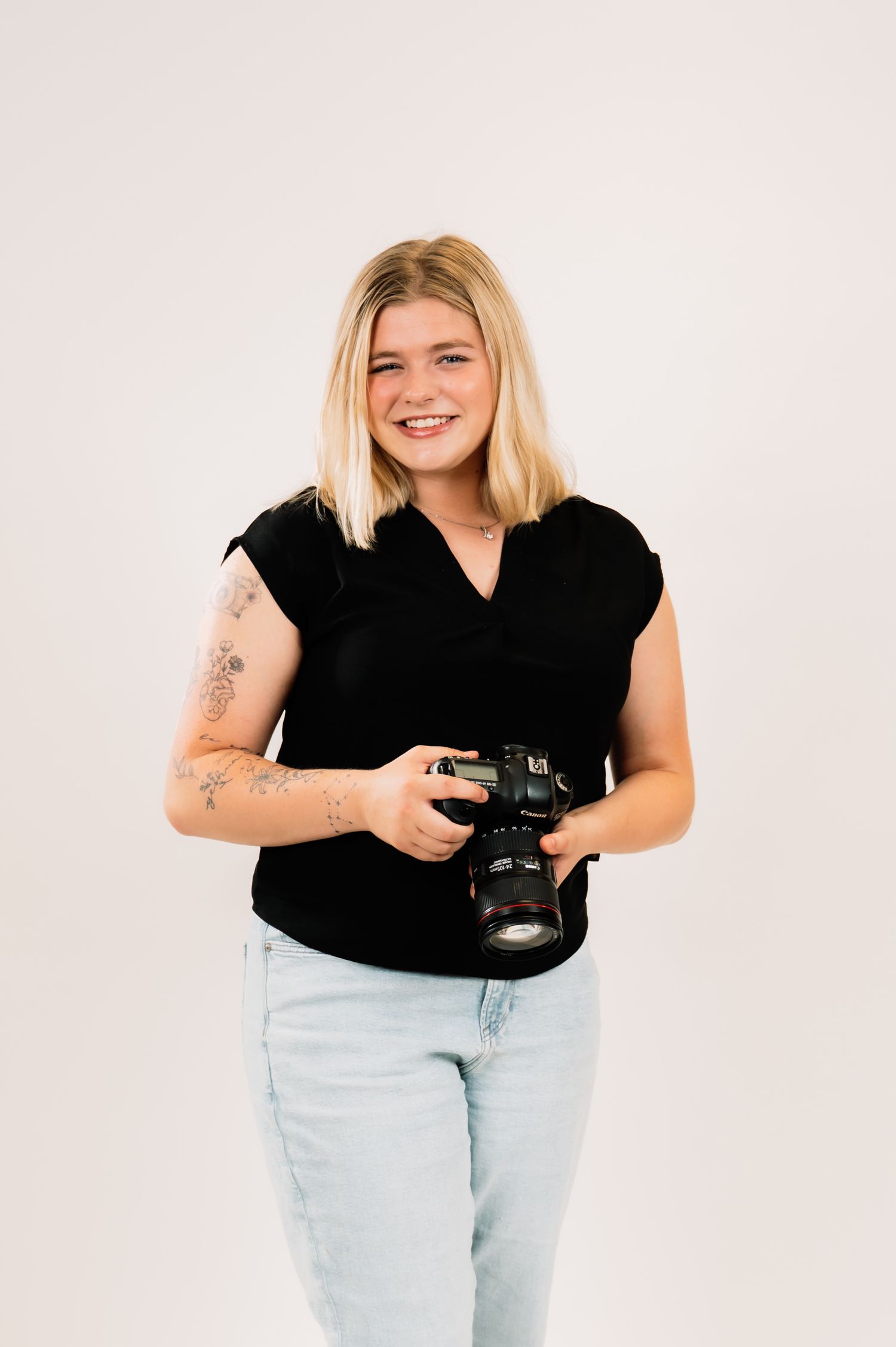 Smiling blonde female photographer standing and holding Canon DSLR camera with floral arm tattoos.