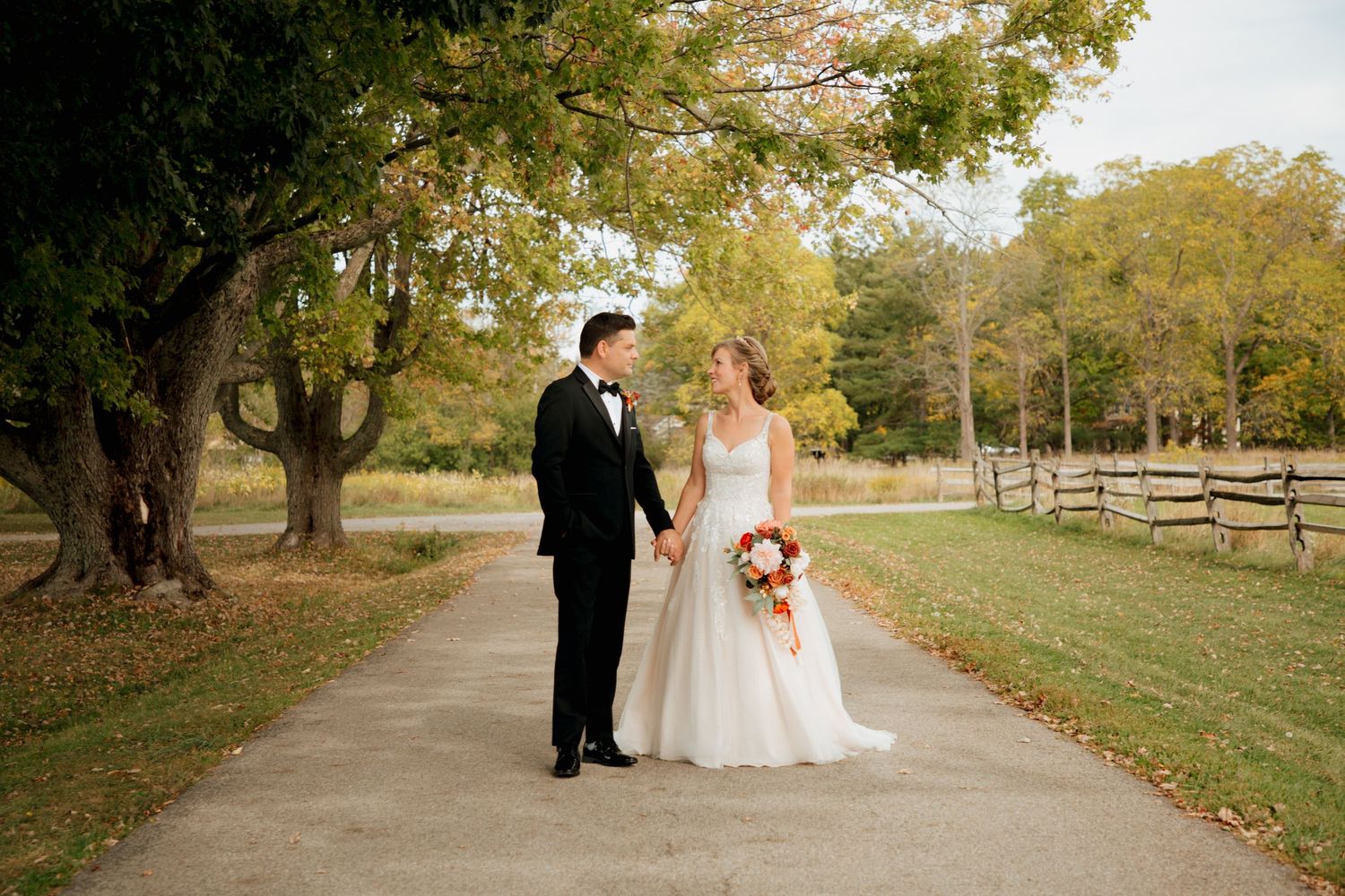 Newlyweds hold hands on rustic tree-lined path with wooden fence and fall scenery.