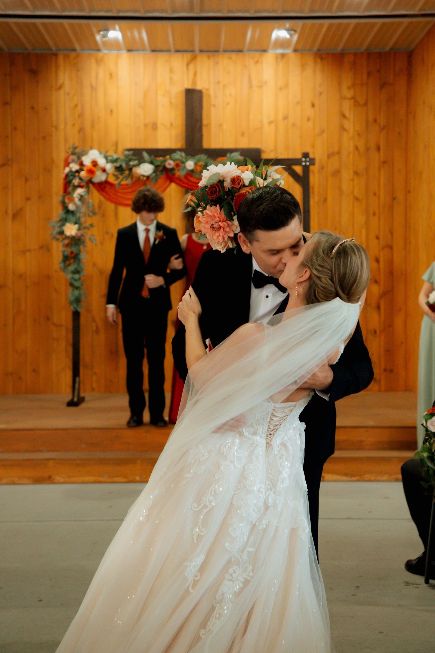 Bride and groom share their first kiss at the altar in a rustic wood-paneled chapel decorated with floral arrangements.