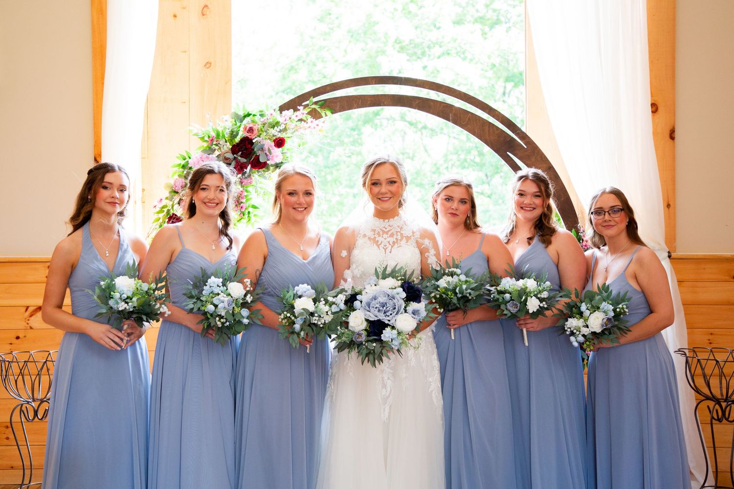 Bride with five bridesmaids in dusty blue gowns holding bouquets under circular arch in barn venue.