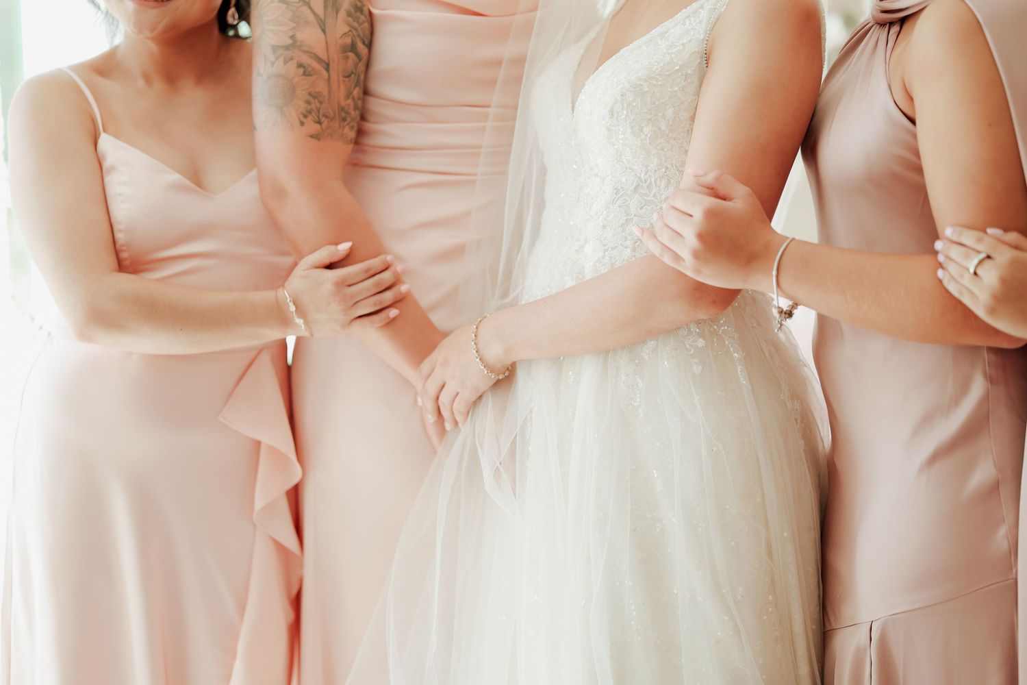 Bride in lace wedding gown standing between two bridesmaids in blush pink dresses holding hands.