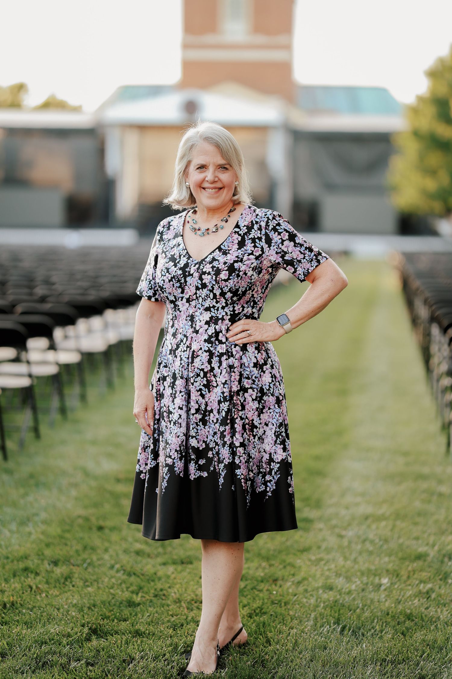 Elegant woman in black floral dress posing confidently on green lawn between rows of black chairs.