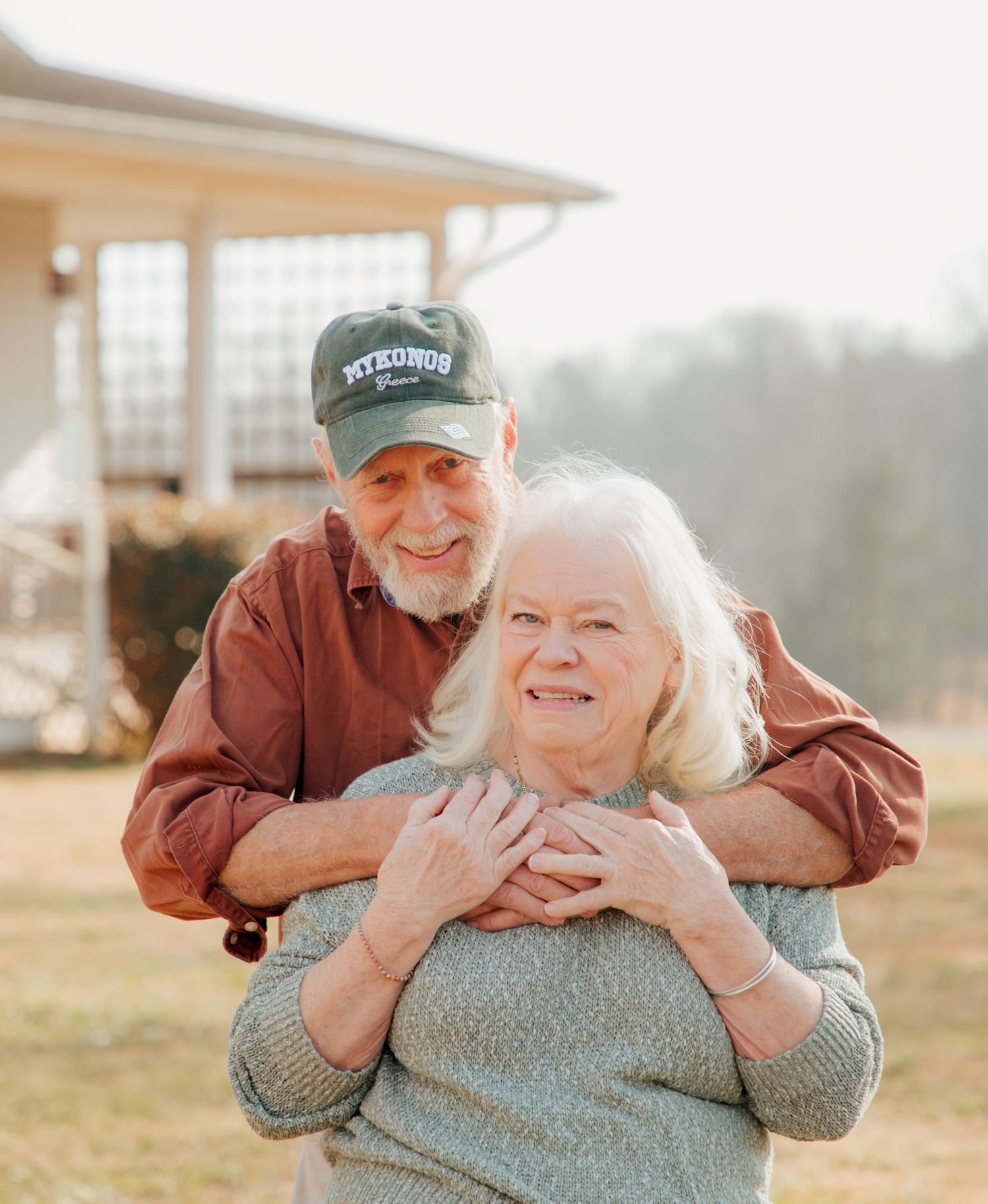 Smiling elderly man embracing woman from behind outdoors, both looking happy and content.