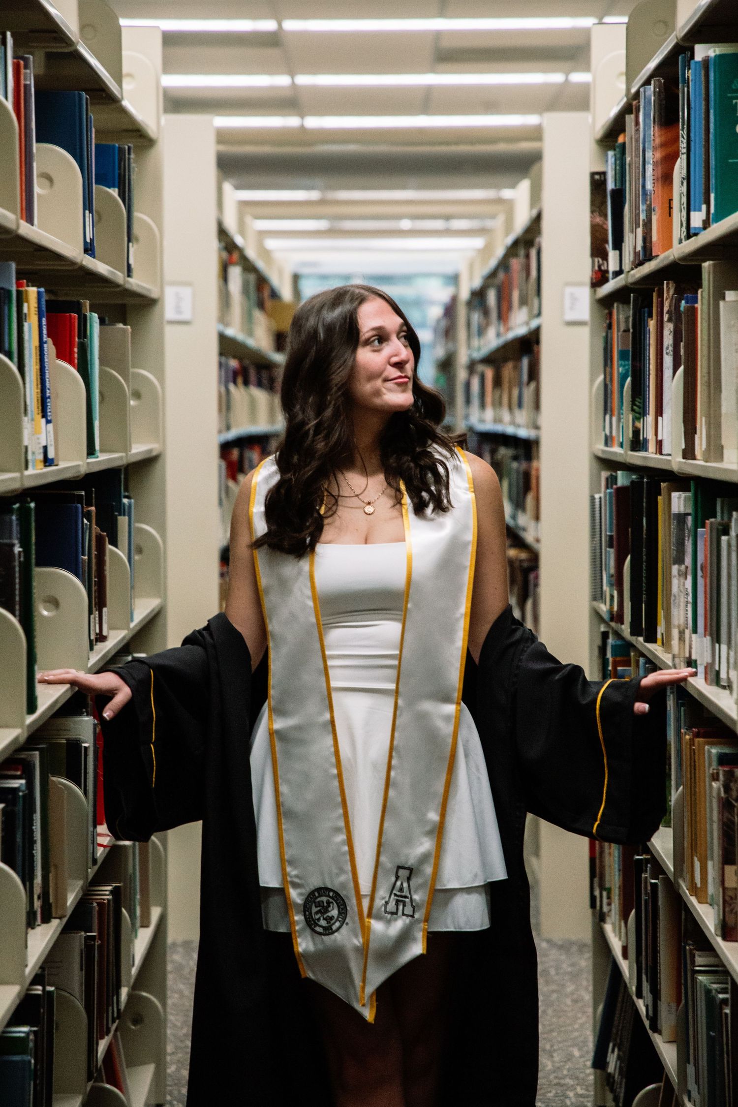 Female graduate in black gown and white stole poses with open arms between library bookshelves.