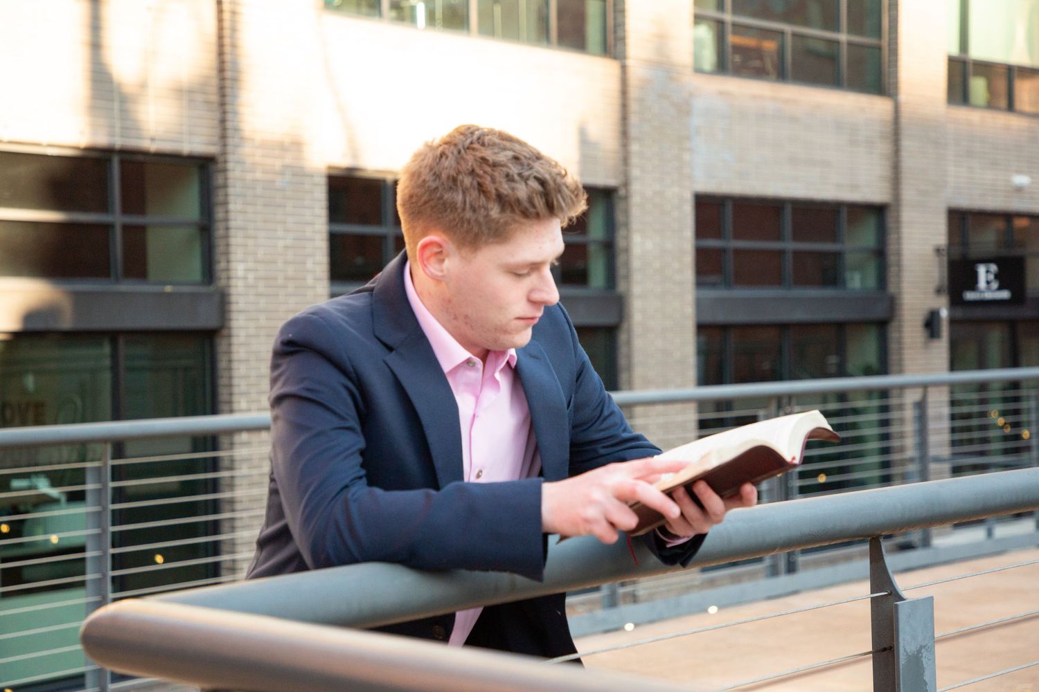 Young man in navy suit and pink shirt reading a book while leaning on an outdoor railing.