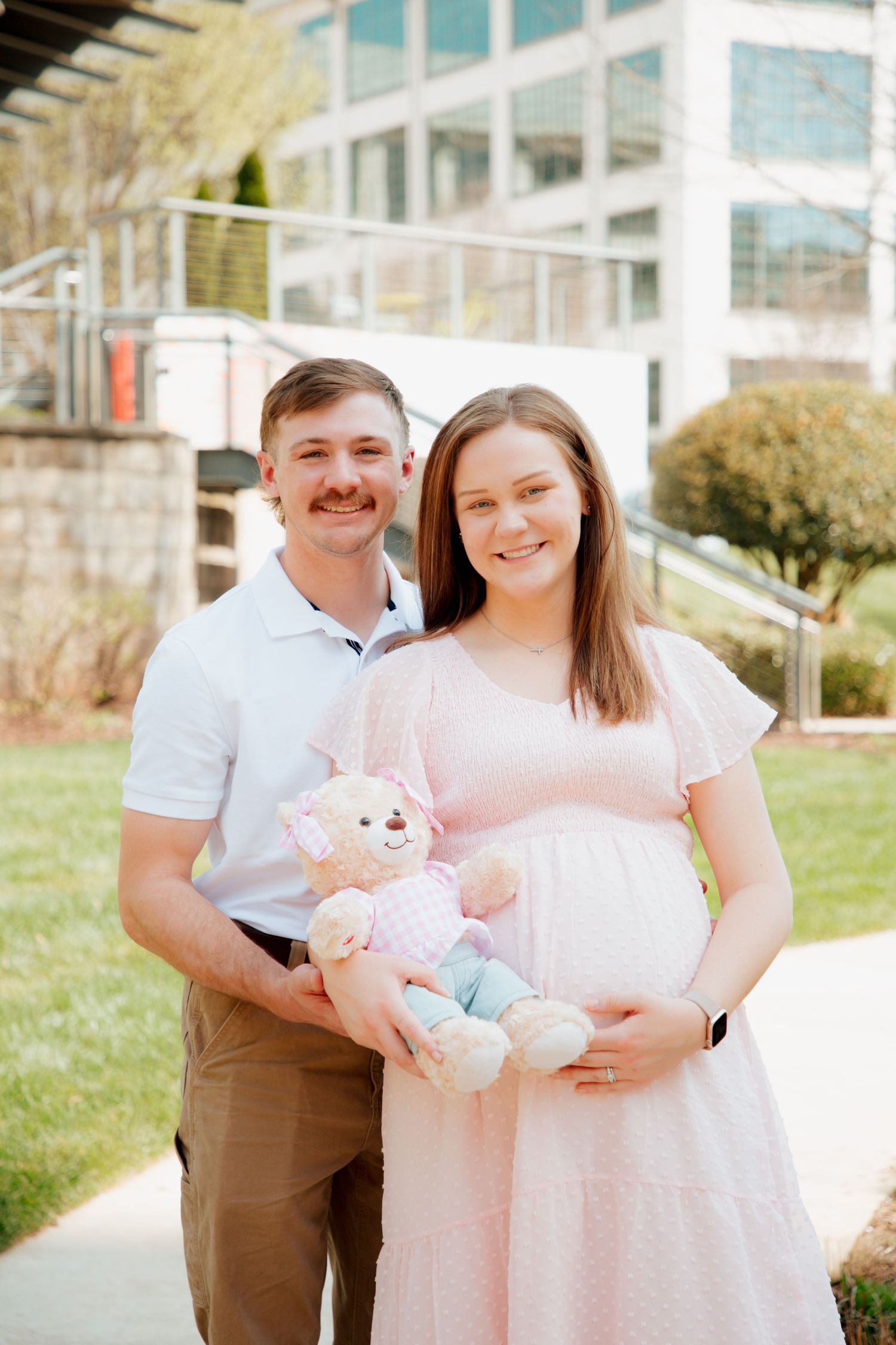 Smiling couple poses outdoors, pregnant woman in pink dress holding teddy bear with partner in white polo shirt.