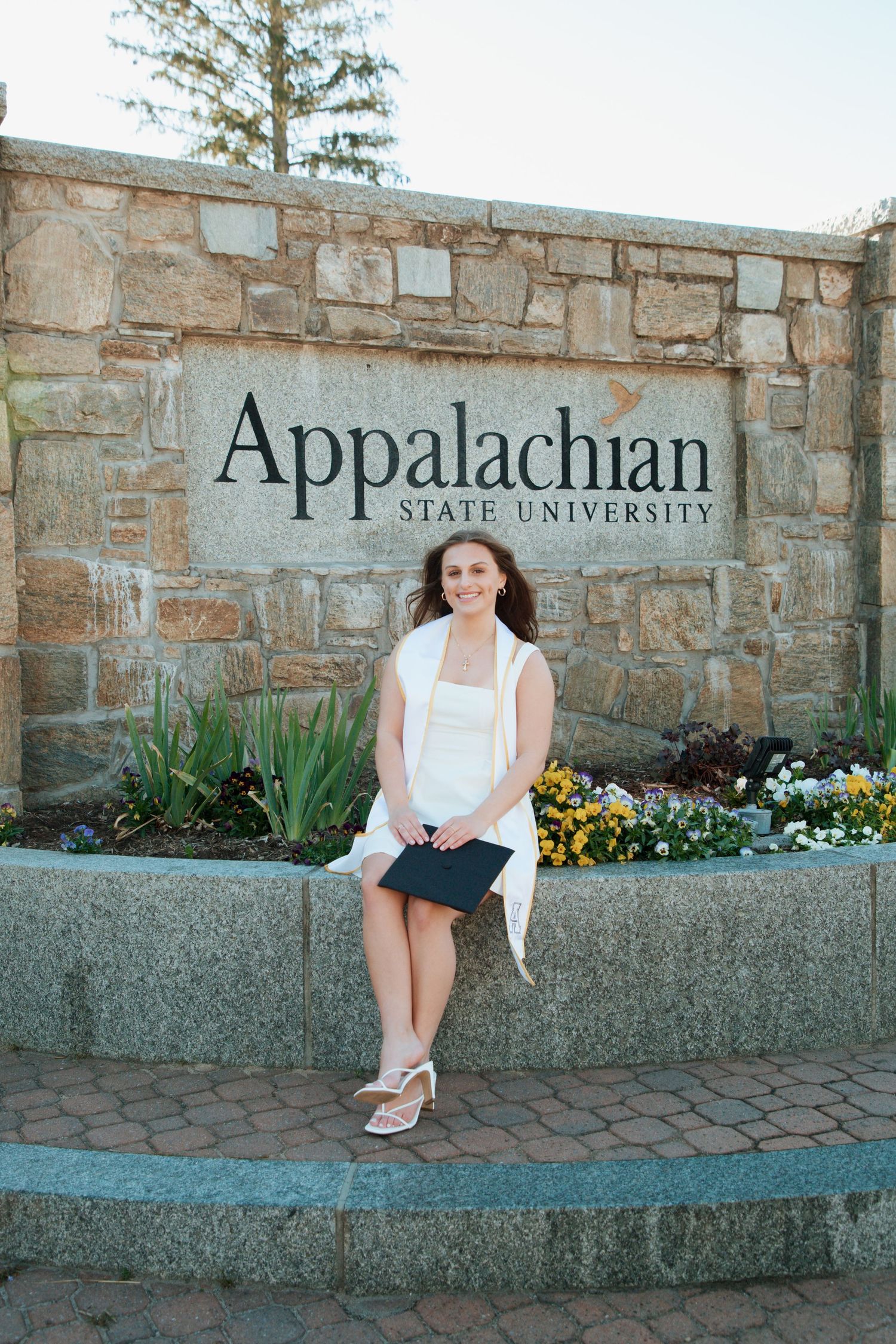 Graduate in white dress sitting at Appalachian State University stone monument sign.