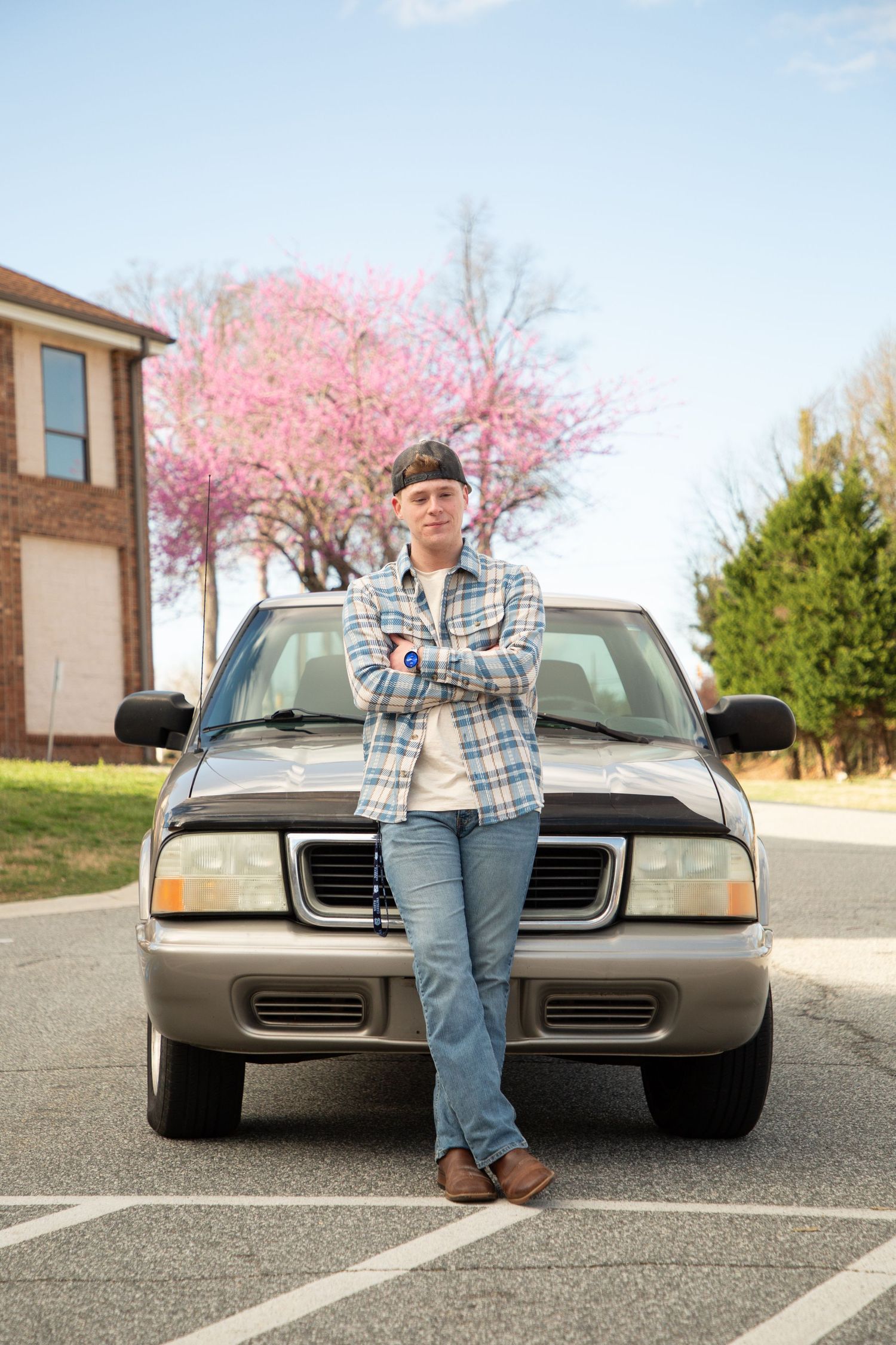 Young man in plaid shirt and cap leaning against a car in a suburban driveway with pink flowering tree.