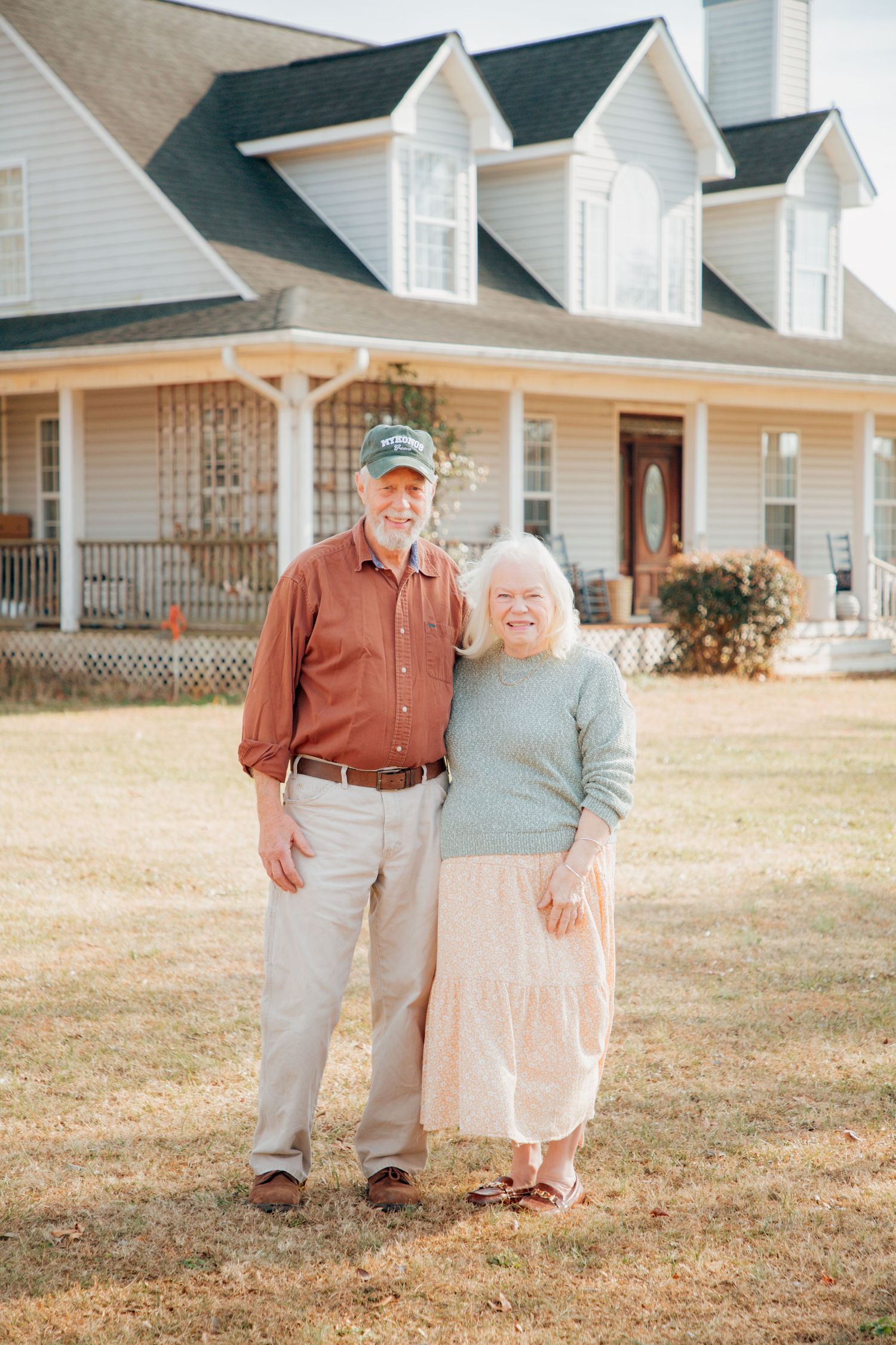 Smiling elderly couple standing together in front of a charming white farmhouse with a wrap-around porch.