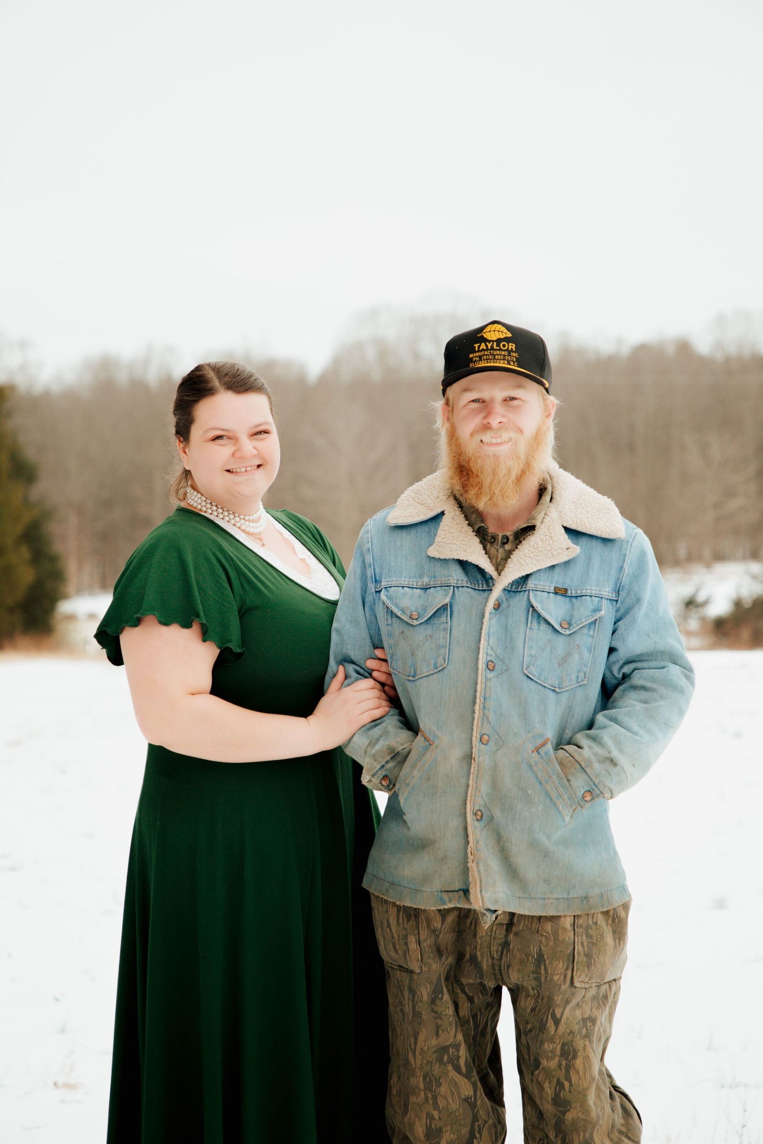 Smiling couple posing together in snowy field, woman in green dress with pearl necklace.