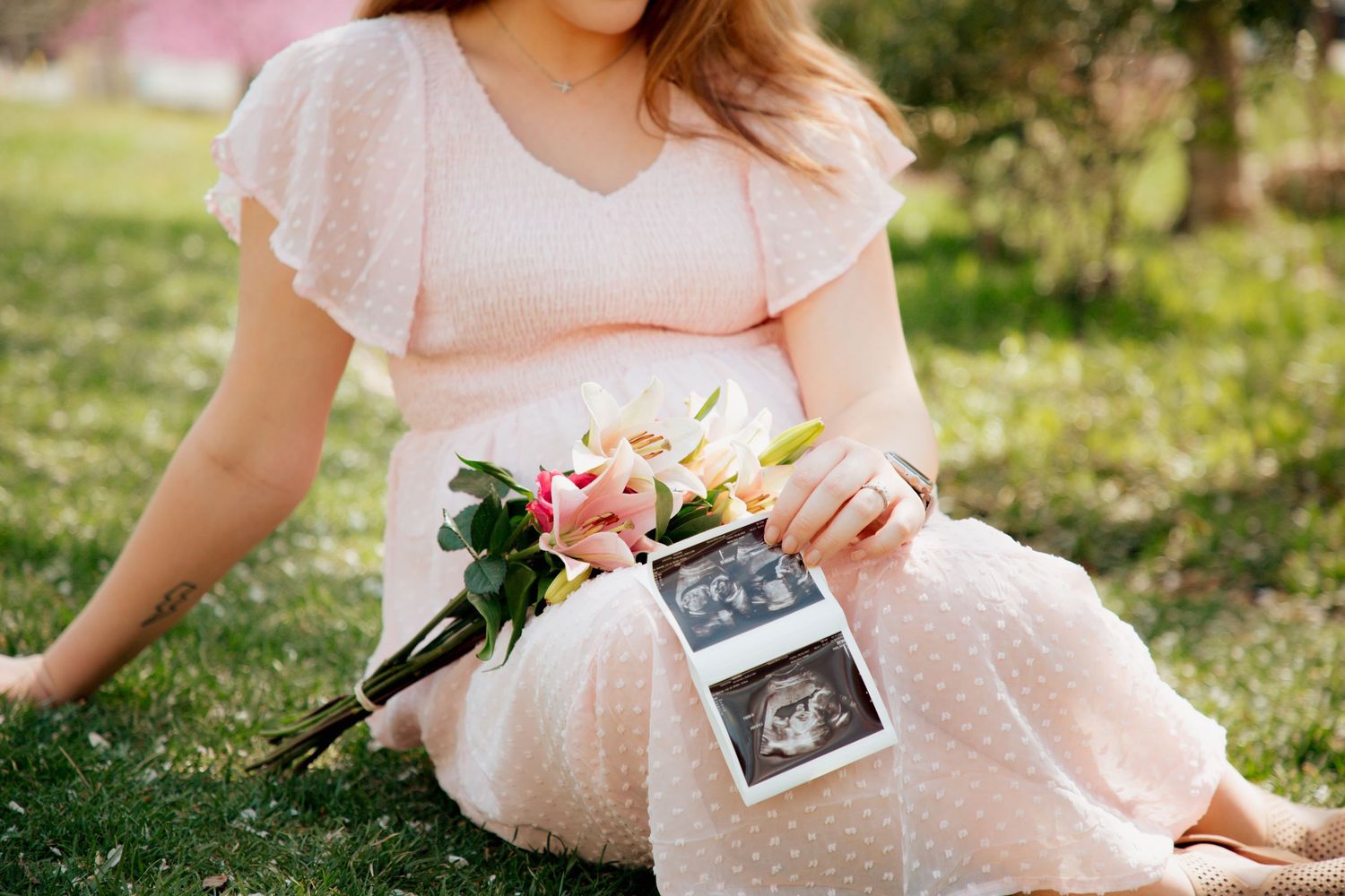 Close-up of pregnant woman in pink polka dot dress holding flowers and ultrasound images.