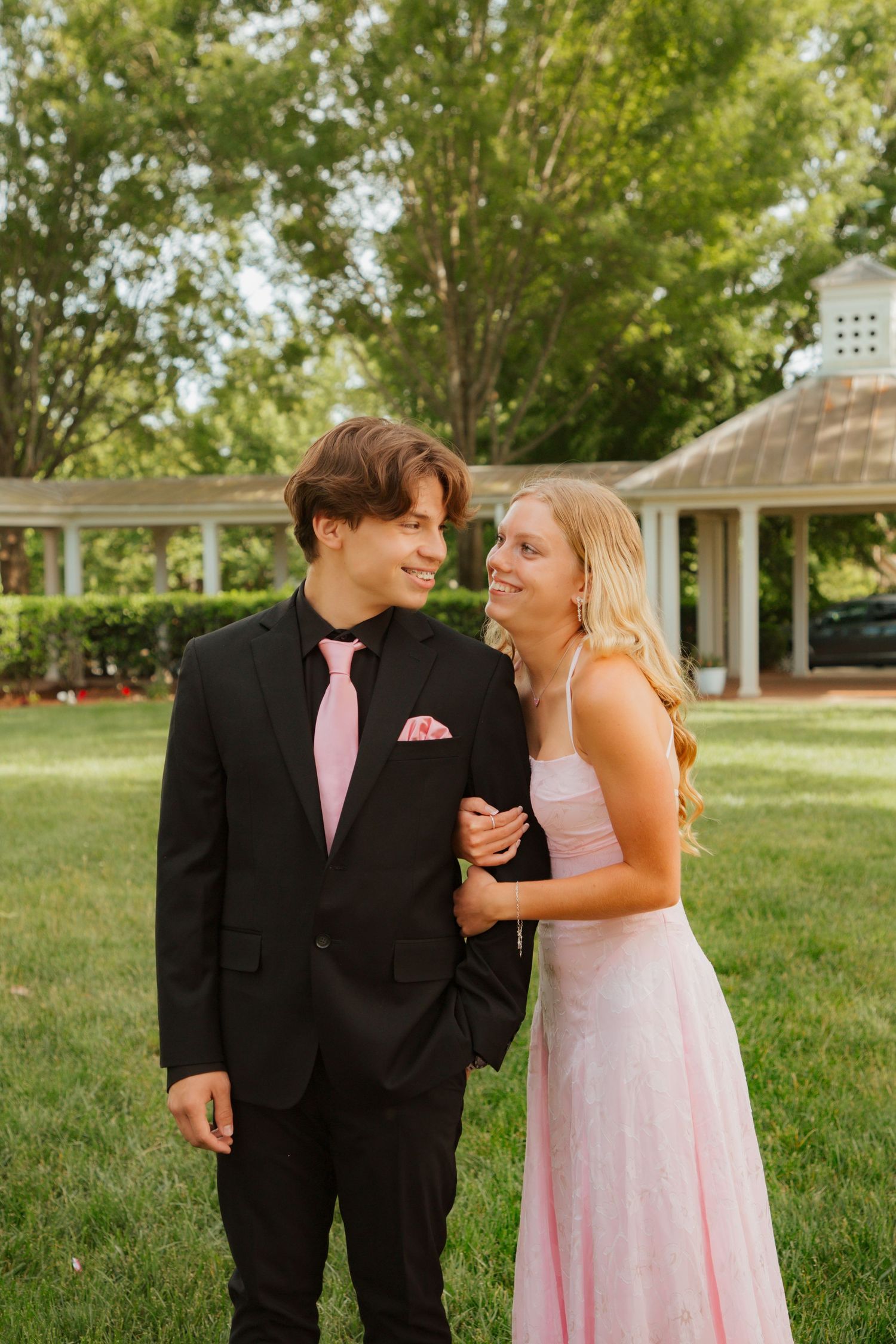 Young couple in prom attire smiling at each other outdoors in a park.