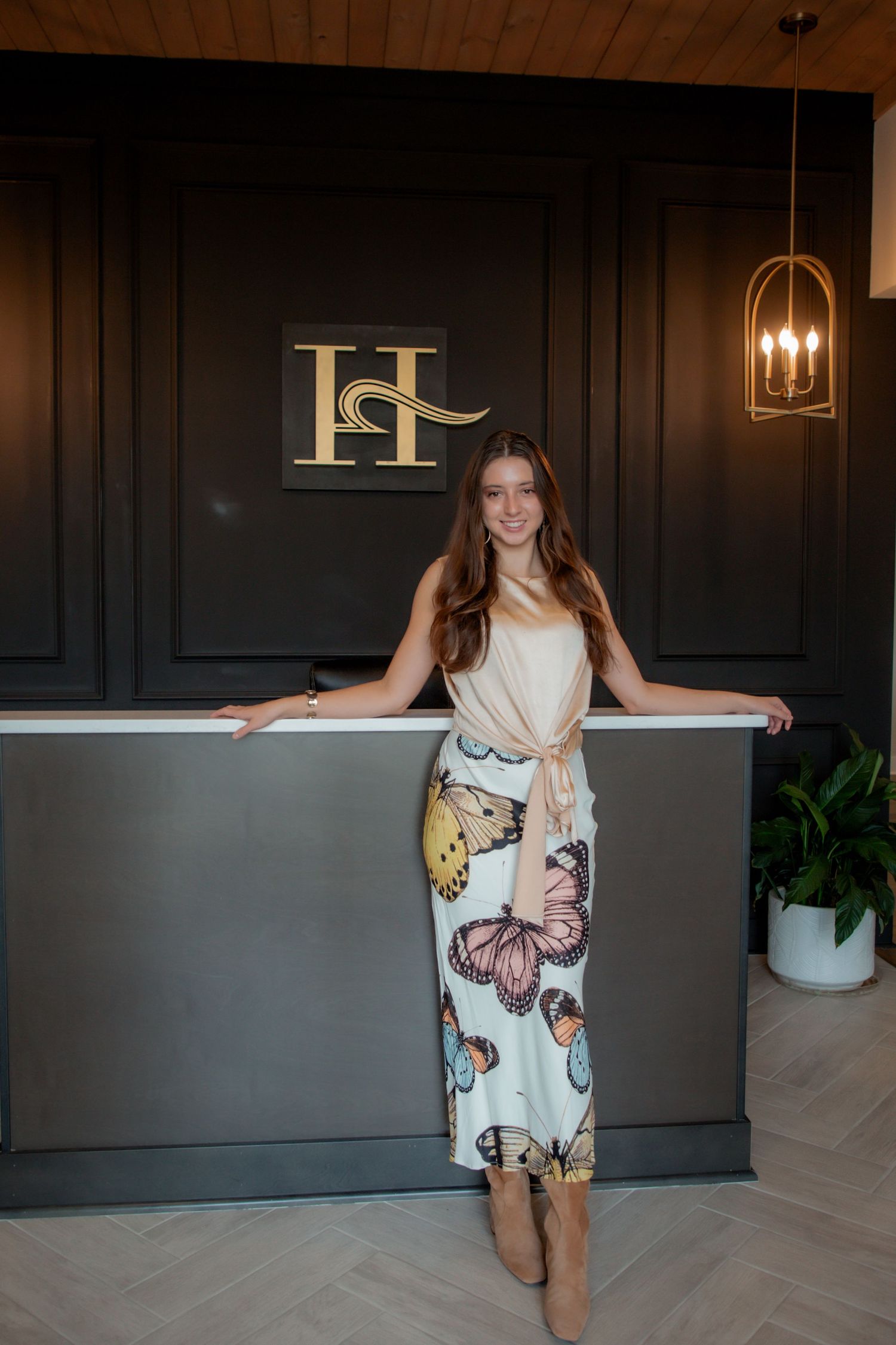 Young woman in butterfly maxi skirt standing at dark reception desk with gold H logo and chandelier.