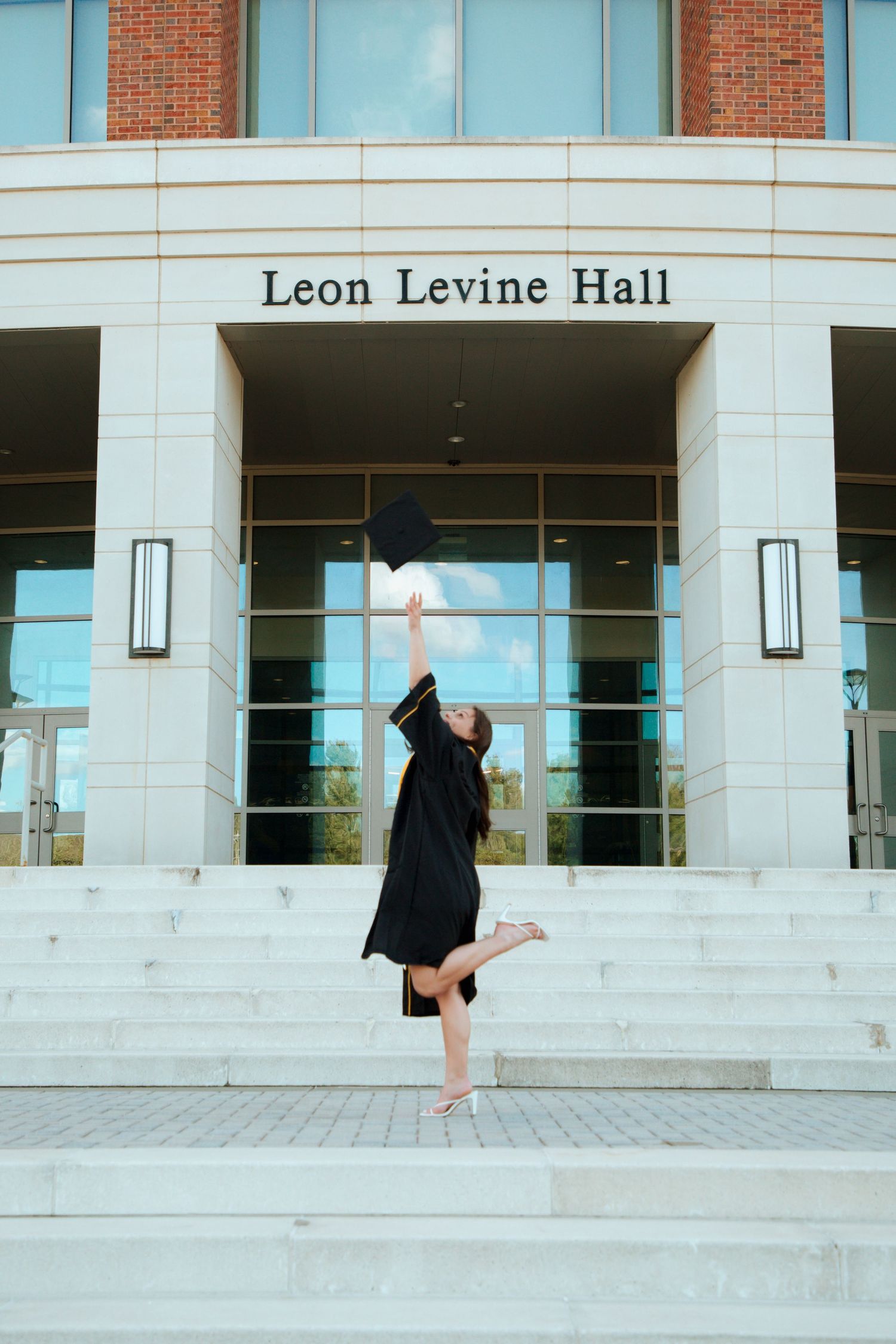 Graduate in black gown tosses cap in air outside Leon Levine Hall building.