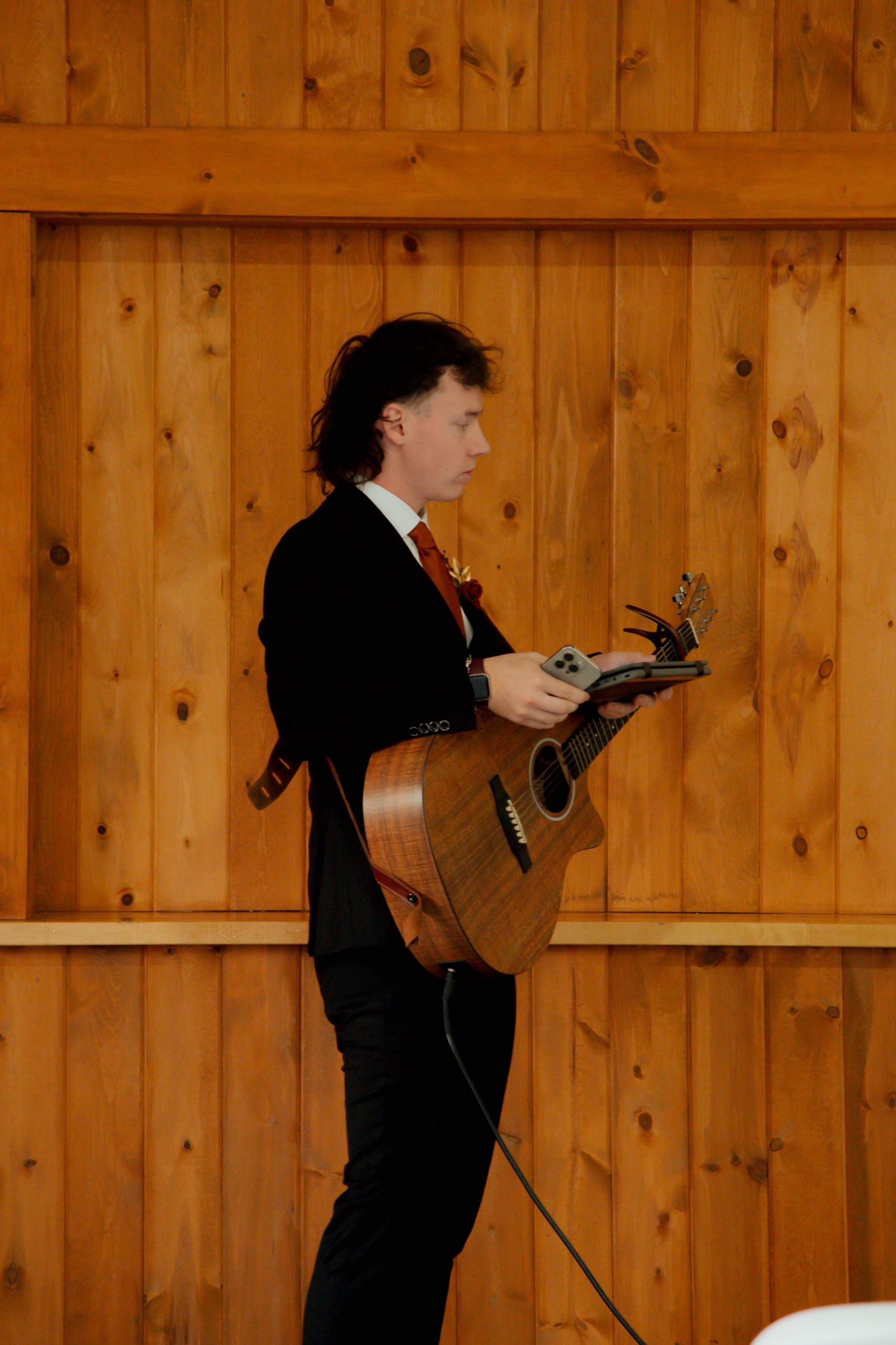 Musician in black suit plays acoustic guitar at rustic wood-paneled wedding venue.