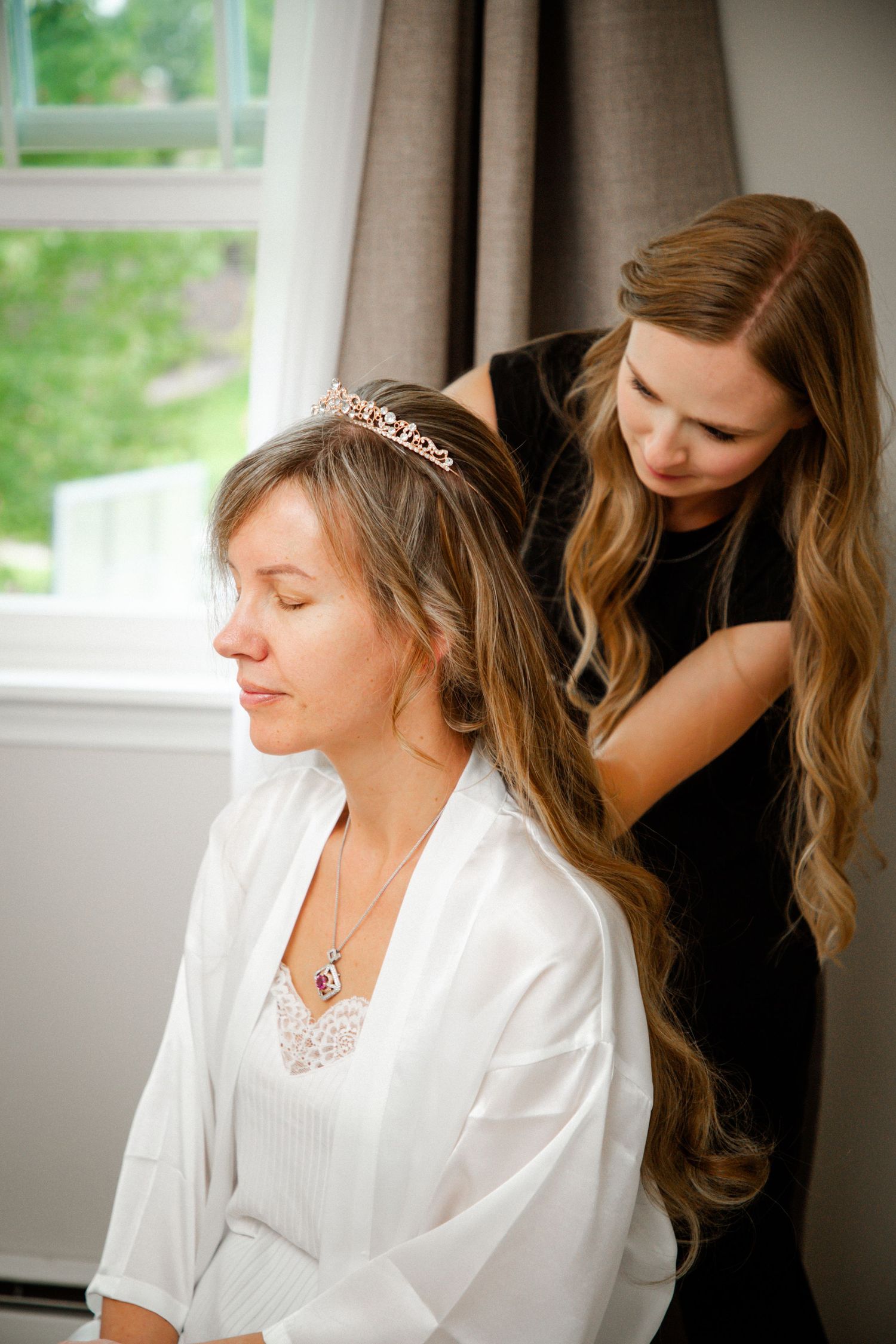 Hair stylist in black dress arranging bride's long hair with tiara during wedding preparation.