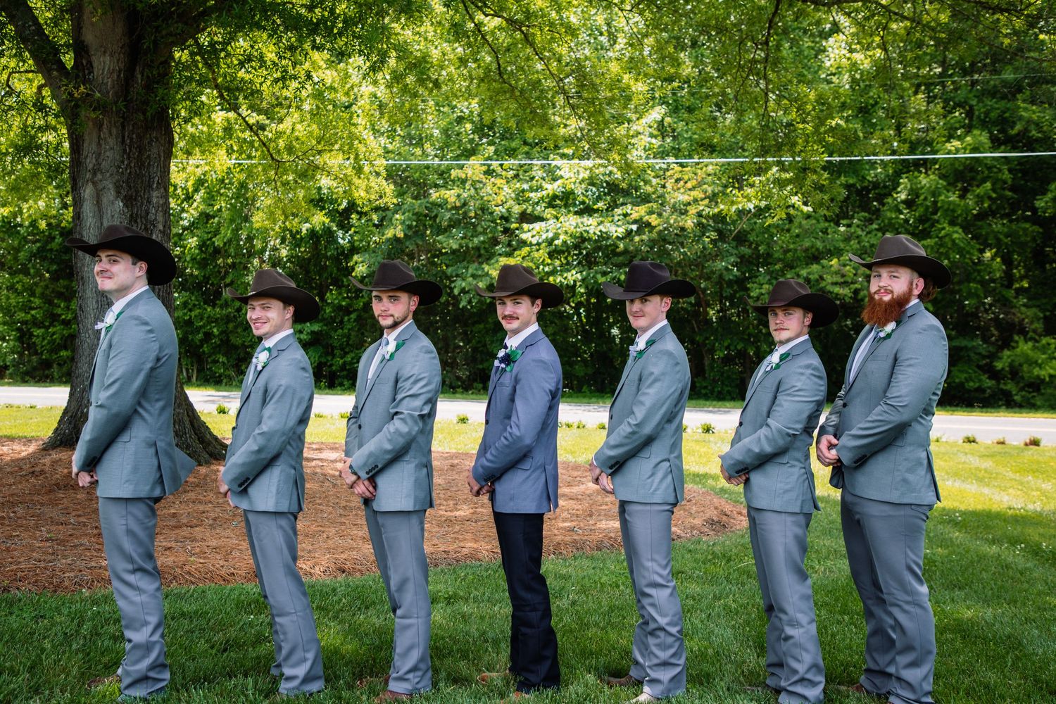 Groomsmen in gray suits and black cowboy hats stand in a side profile line outdoors at a country wedding.