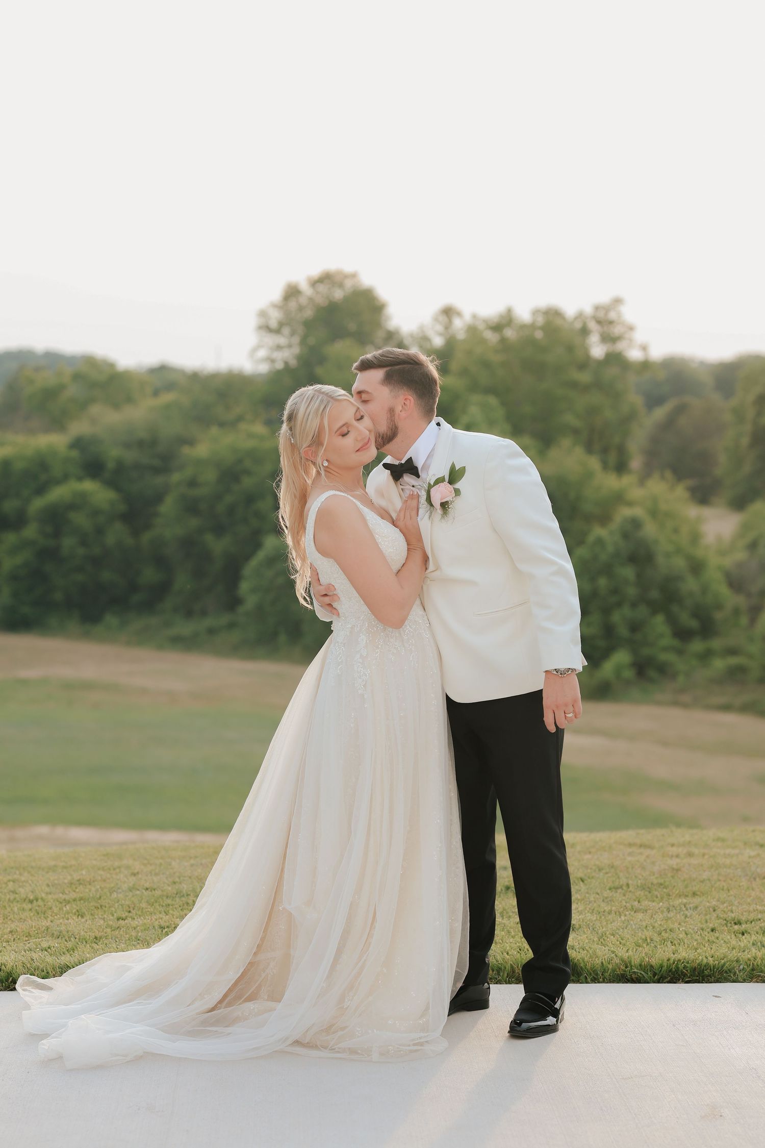 Groom in white tuxedo kisses bride in flowing gown at sunset outdoor wedding portrait.