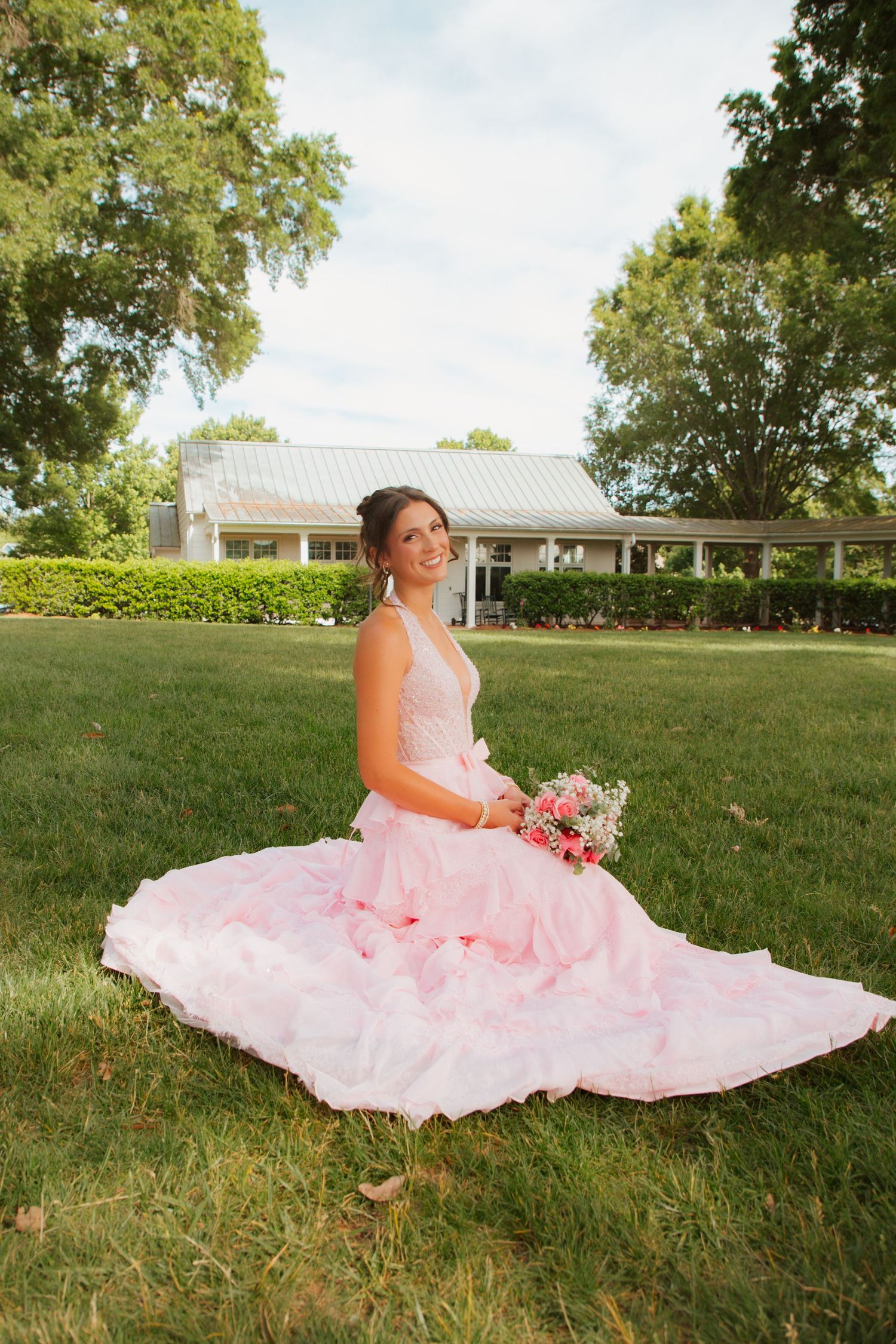 Bride in pink ball gown sitting on green grass holding bouquet, with white house and trees in background.
