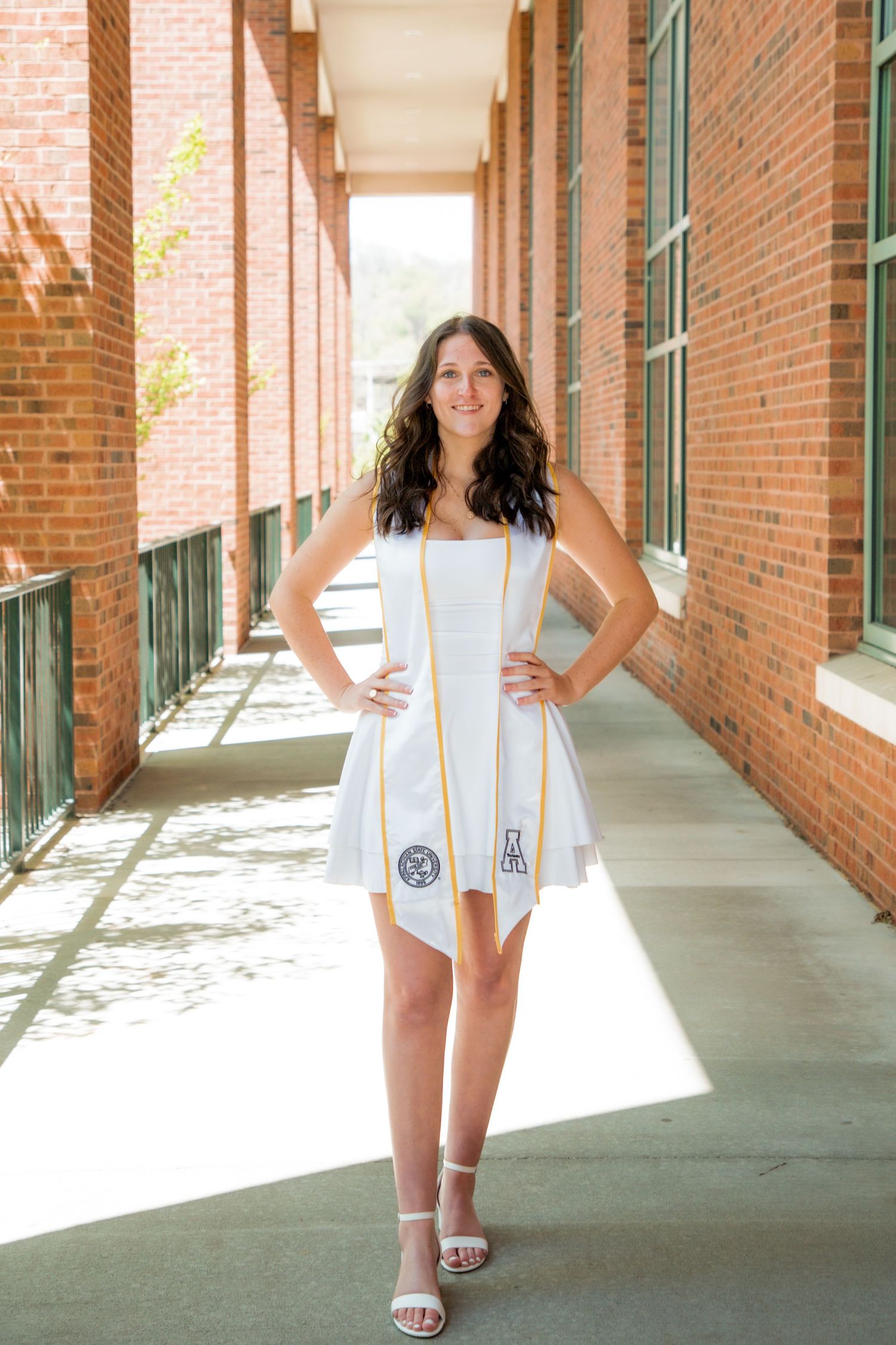 Female graduate in white dress and gold stole posing in brick hallway.