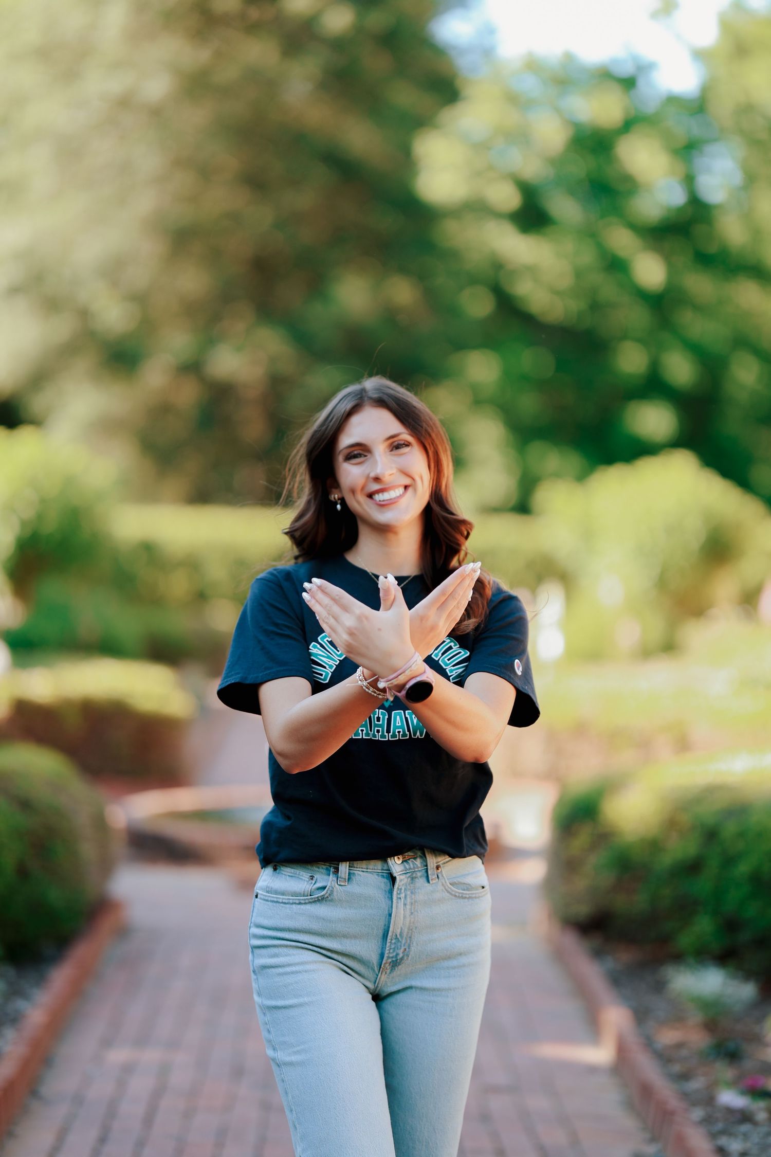 Smiling young woman in UNCW Seahawks shirt making an X gesture with her arms on a brick garden path.