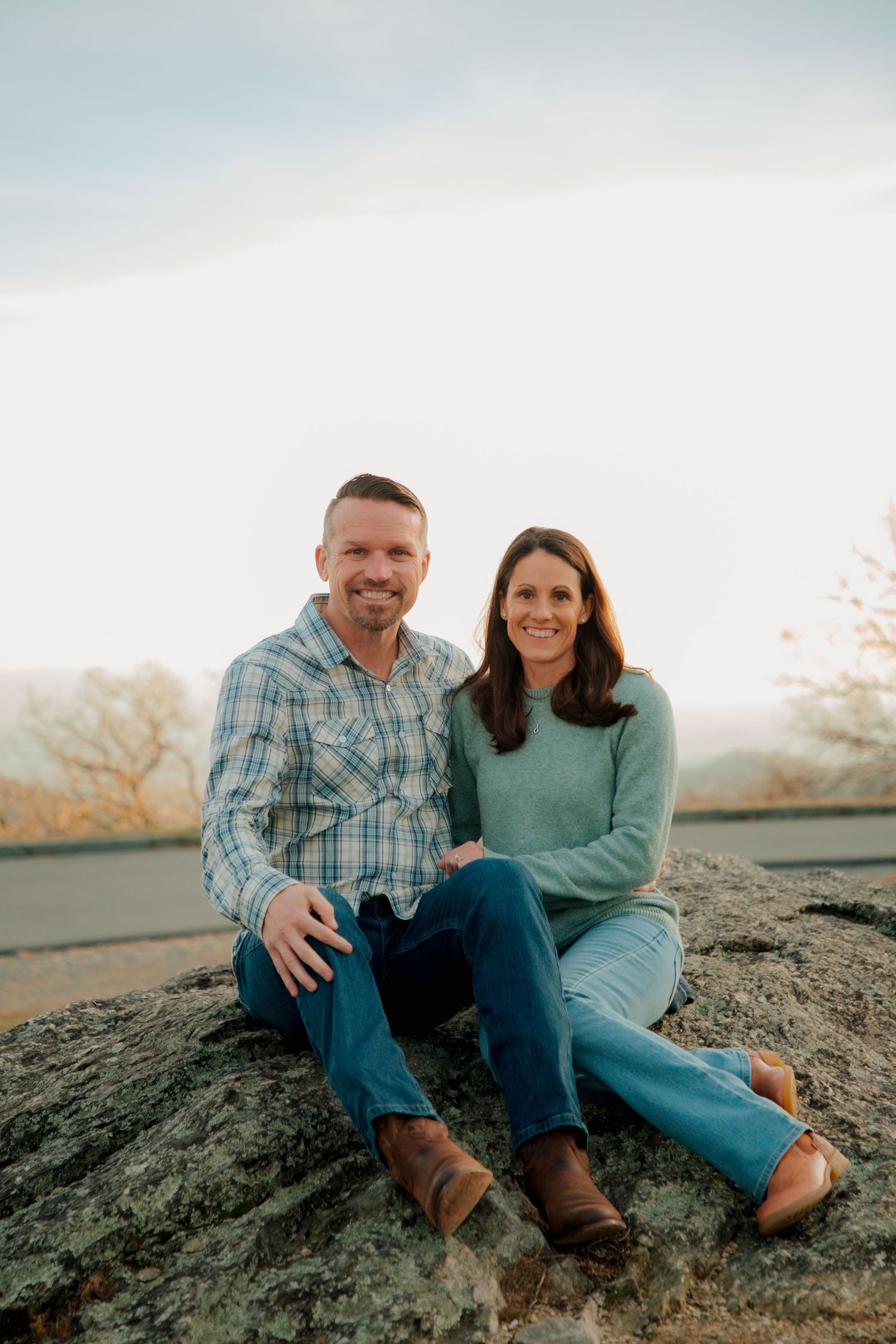 Smiling couple sits together on a large rock with a scenic mountain landscape at sunset.