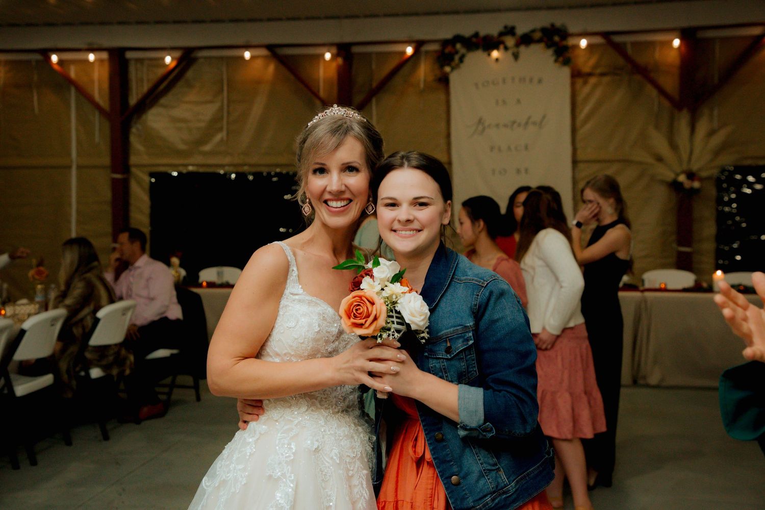 Bride in lace wedding gown posing with guest holding bridal bouquet at reception.