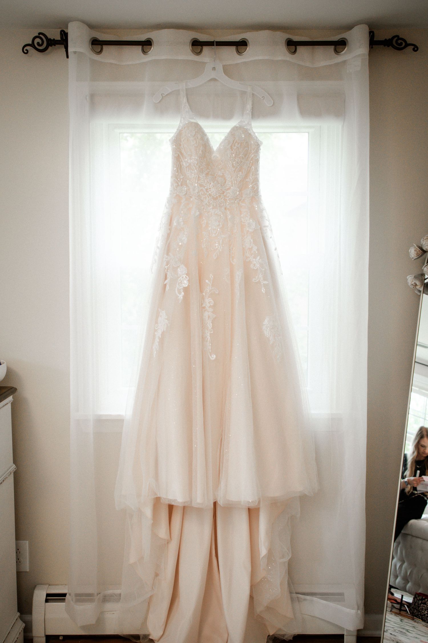 Elegant blush lace A-line wedding dress hanging on curtain rod by window.